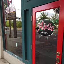 The red and green storefront and window display featuring the logo for Abella Pizzeria, representing a local spot for casual Italian dining, pizzas, and classic favorites.