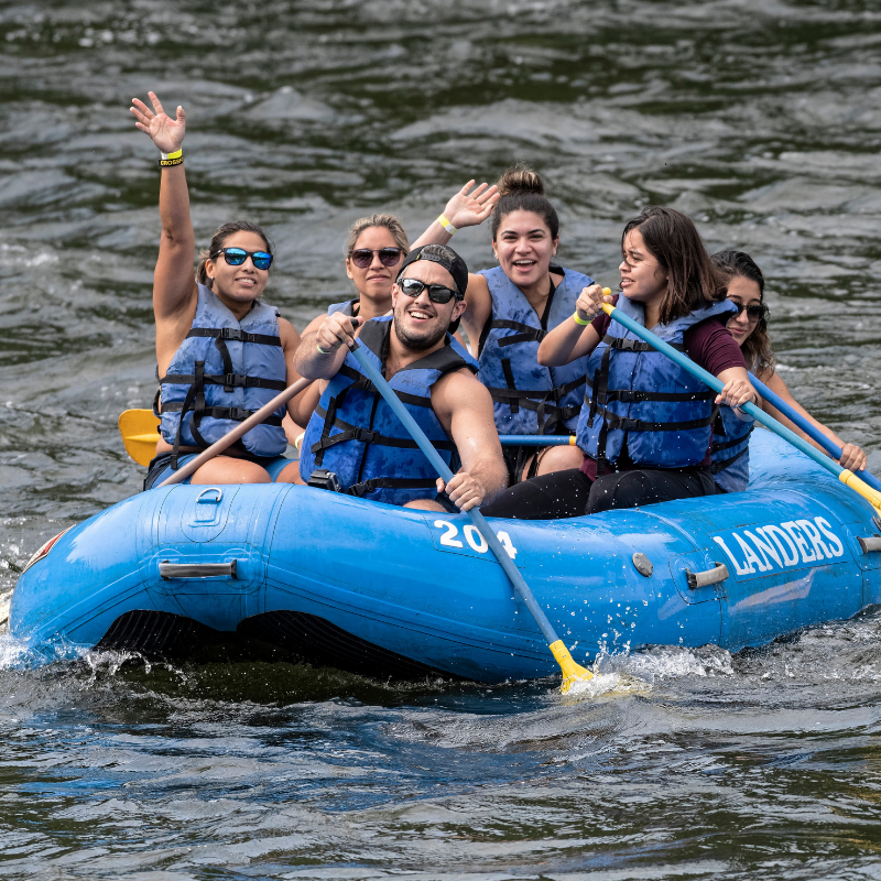 A group of six people smiling and paddling together in a blue 'LANDERS' raft on the river, wearing life vests, highlighting the exciting river rafting and summer adventure experiences offered by the outfitter.