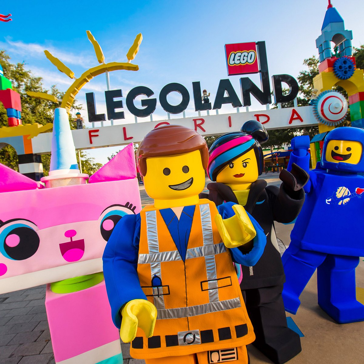 Emmet, Wyldstyle, and Unikitty costumed characters posing cheerfully in front of a colorful LEGO sign, representing the fun, rides, and themed attractions at the LEGOLAND family amusement park.