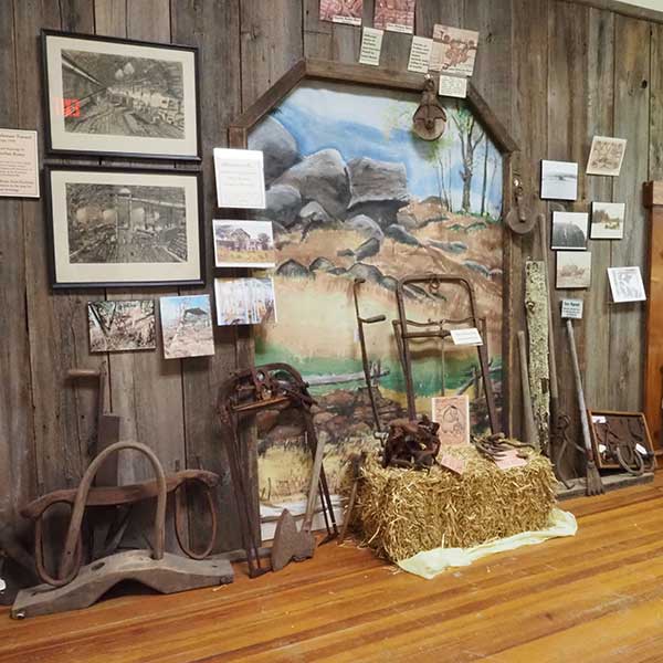 A rustic historical museum display featuring antique farm implements, a bale of straw, framed black-and-white drawings of vintage trains, and a central mural, set against a wooden plank wall.