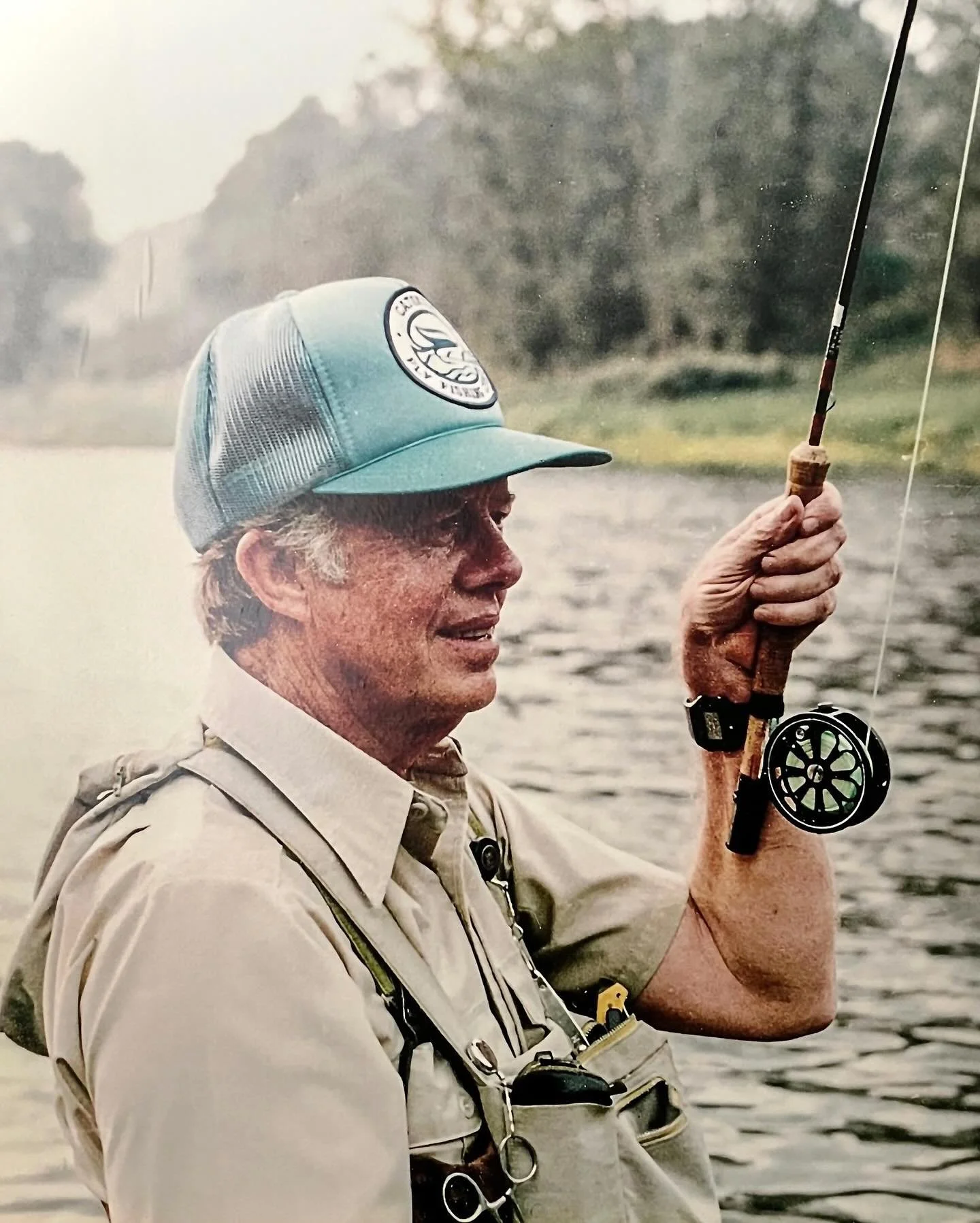 A close-up photograph of an angler fly-fishing in a river, holding a rod and reel while wearing fishing waders, representing the culture and education celebrated by the renowned fly-fishing center.