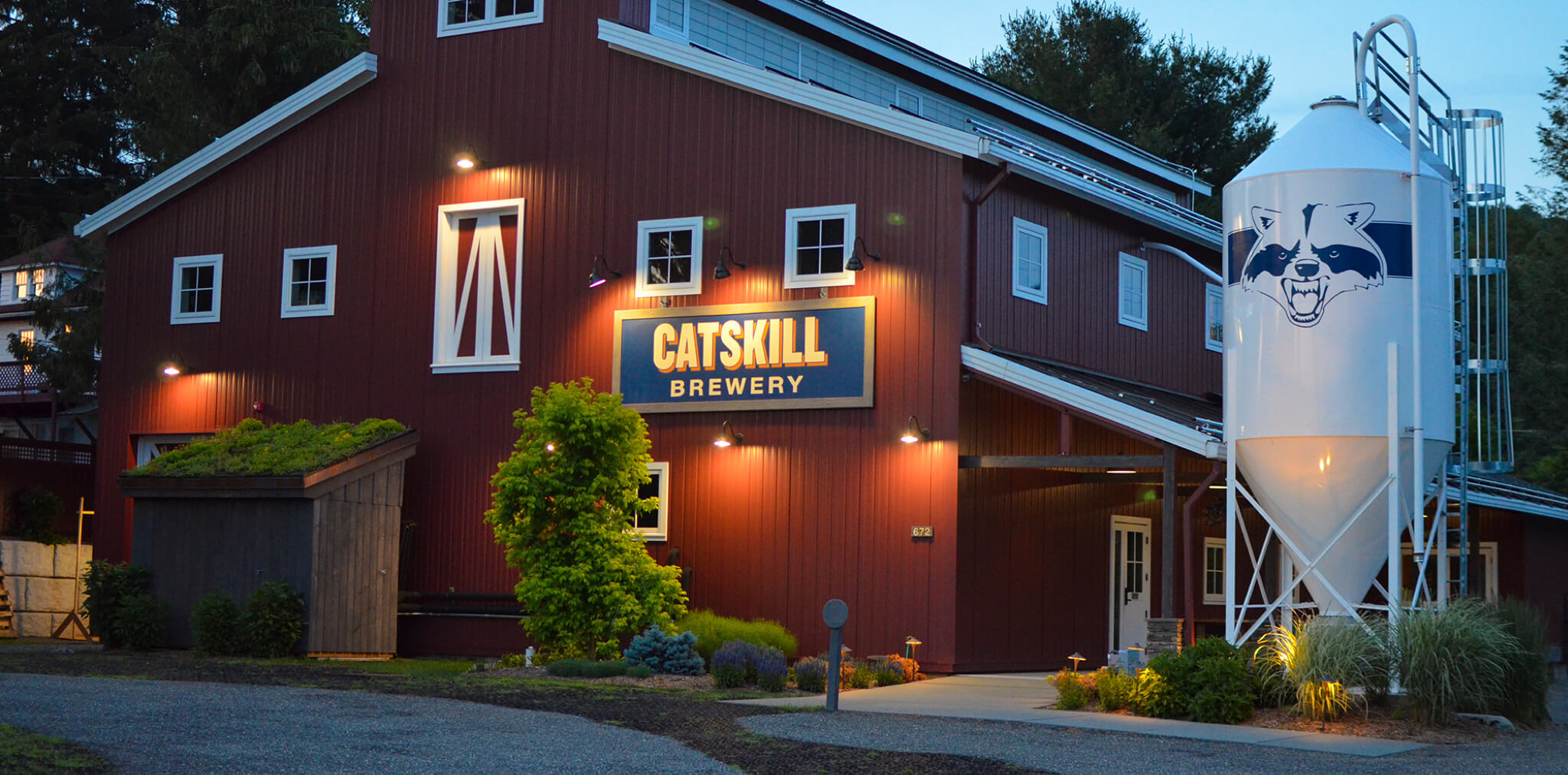 The exterior of the Catskill Brewery, a large red barn-style building illuminated at dusk with a prominent sign and a white silo featuring the raccoon mascot, highlighting the spot for craft beers and the lively tasting room.