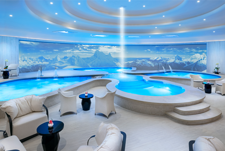 Luxurious and modern indoor spa retreat featuring tiered blue pools, plush white seating, and a large mountain mural backdrop, illustrating a peaceful space for wellness, massages, and relaxation experiences.