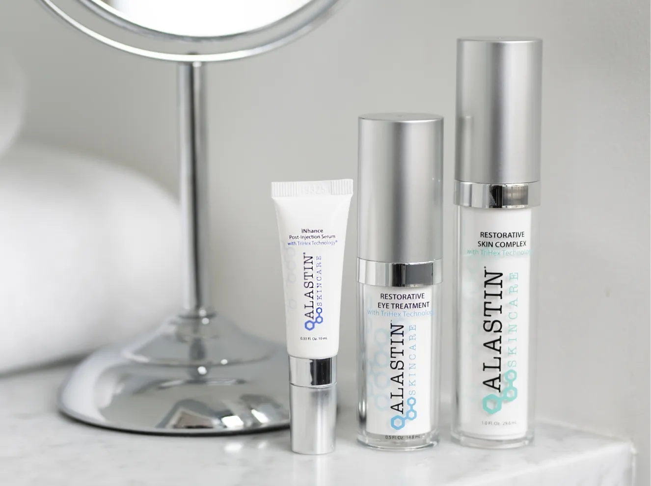 Three Alastin Skincare products: iNhanse Post-Injection Serum, Restorative Eye Treatment, and Restorative Skin Complex, displayed next to a mirror on a white countertop. at Becker Cosmetic featuring cosmetic treatment services