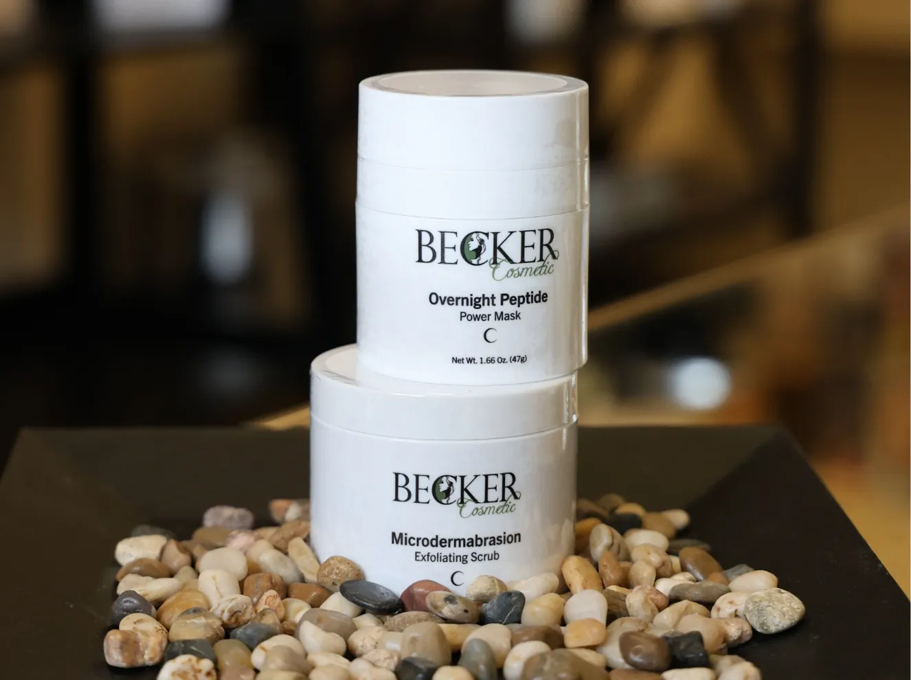Two Becker Cosmetic skincare products: an 'Overnight Peptide Power Mask' and a 'Microdermabrasion Exfoliating Scrub', displayed on a bed of decorative pebbles. at Becker Cosmetic featuring Skincare Products services
