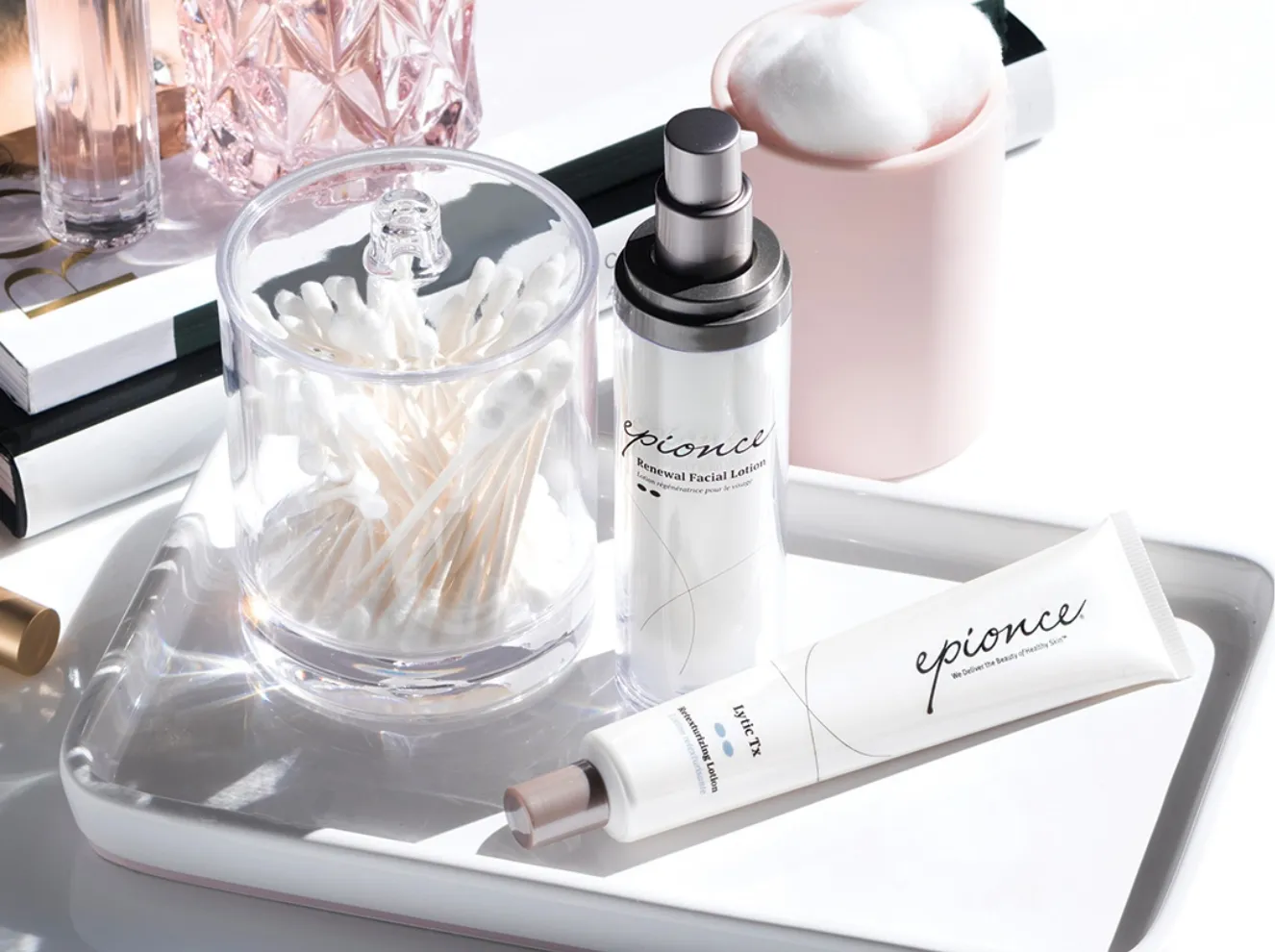 Arrangement of Epionce Renewal Facial Lotion, Lytic Tx Retexturizing Lotion, cotton swabs, and a pink mug with bath puffs on a white tray, showcasing skincare products. at Becker Cosmetic featuring Medical-Grade Skincare services