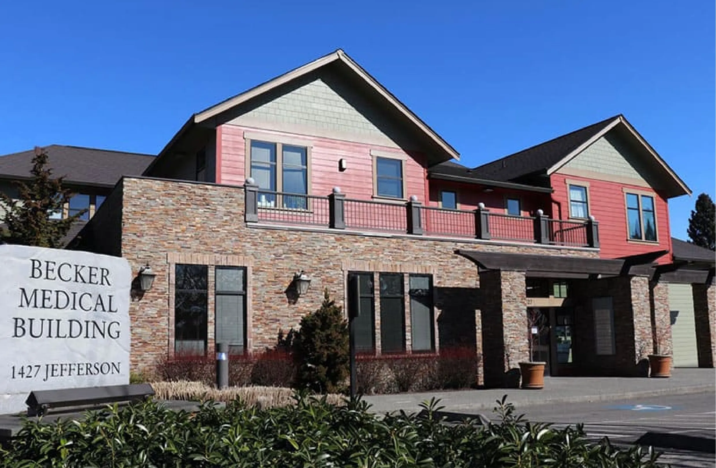 Exterior view of the Becker Medical Building, a multi-story building with red siding and natural stone accents, visible under a clear blue sky. A sign for 'Becker Medical Building, 1427 Jefferson' is in the foreground. at Becker Cosmetic cosmetic clinic in Enumclaw WA
