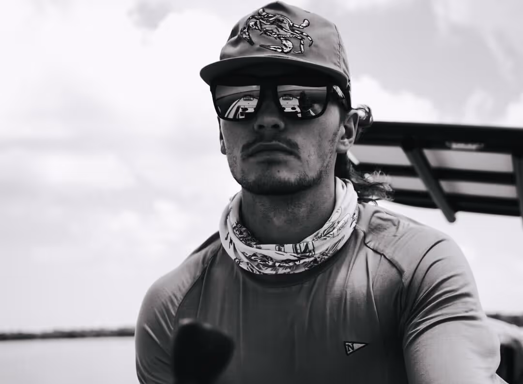 Man wearing sunglasses, a cap, and a neck gaiter sitting on a boat with water and sky in the background.