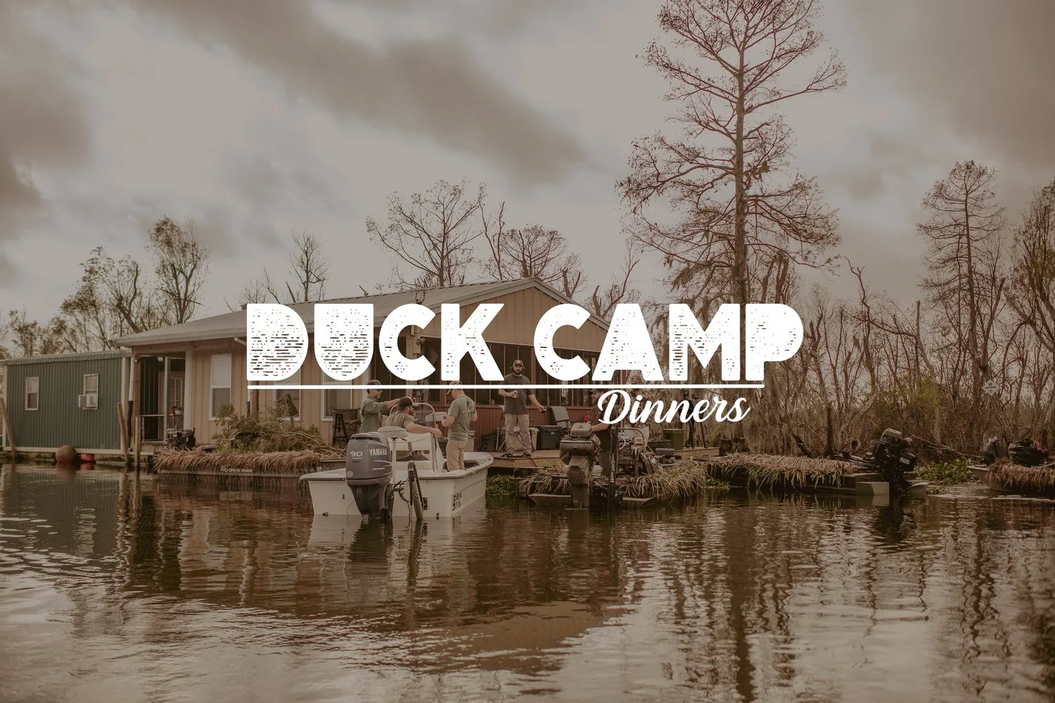Group of people in a small boat near a dock with houses and bare trees reflected in calm water under a cloudy sky.
