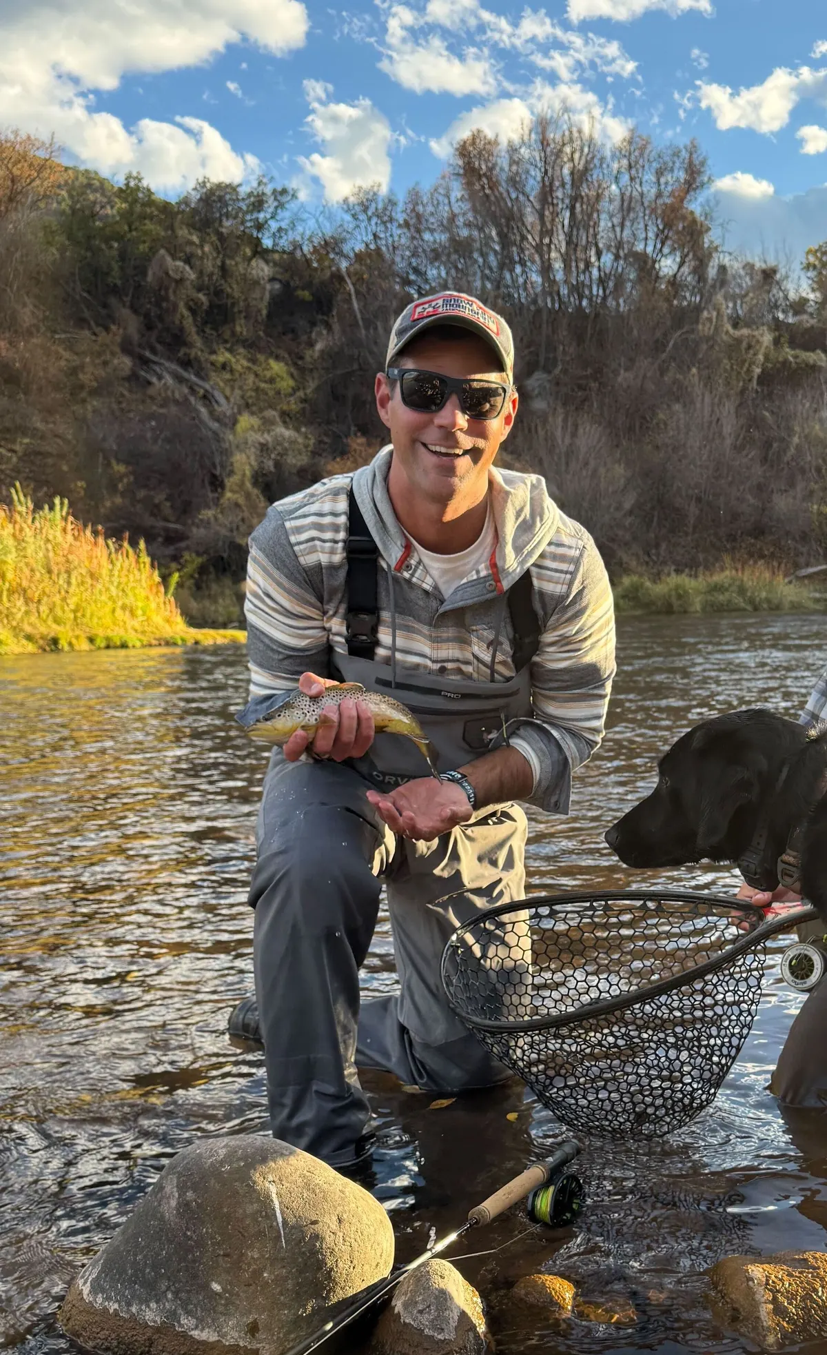 Man wearing camouflage clothing and green cap holding a shotgun in tall dry reeds.