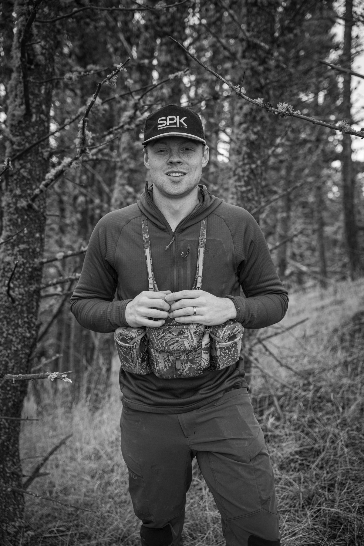 Man in camouflage clothing and green cap holding a camouflaged shotgun in a field of dry tall grass.