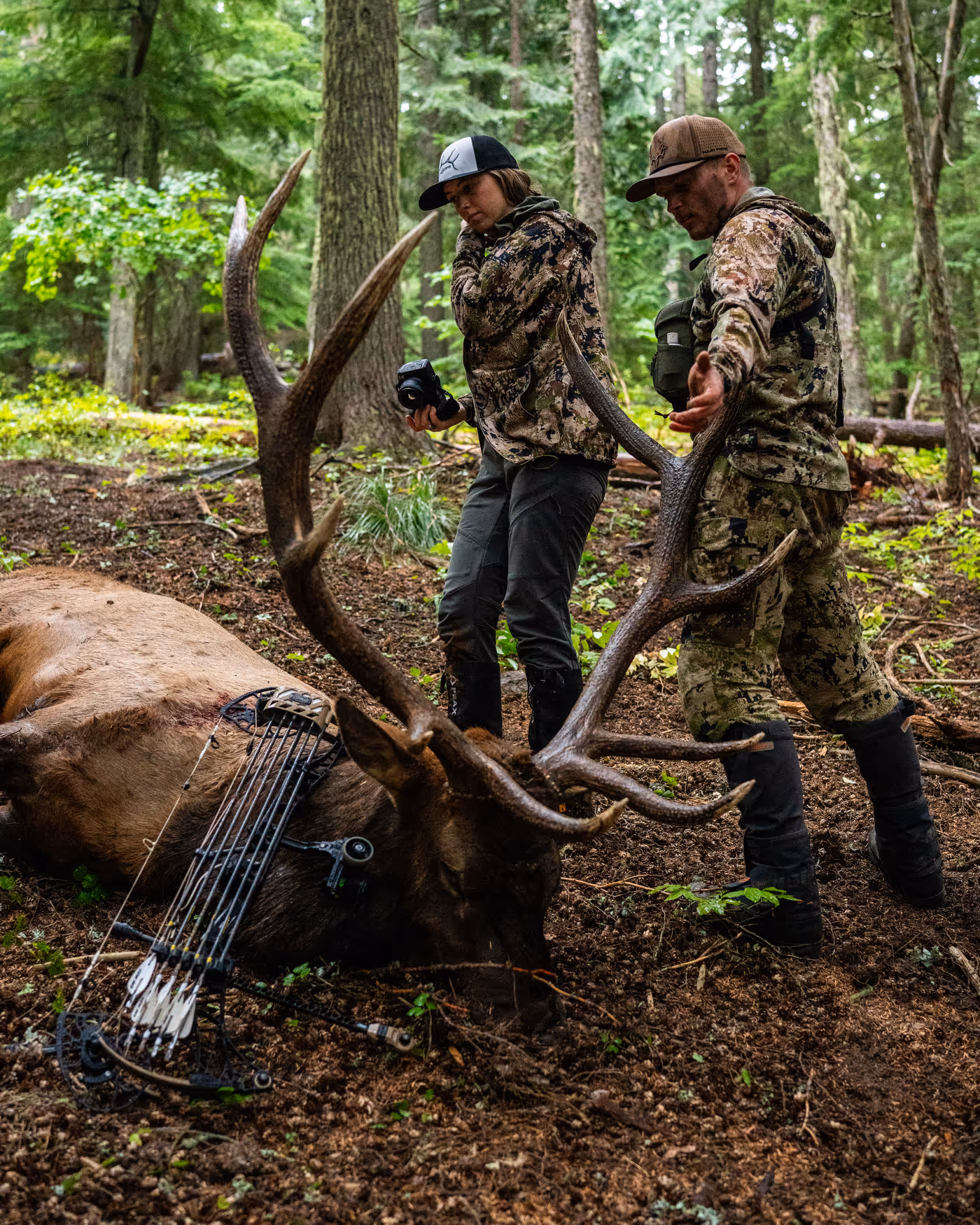 Two people wearing camouflage gear stand back-to-back in shallow water, one holding a fishing rod and the other holding a rifle.