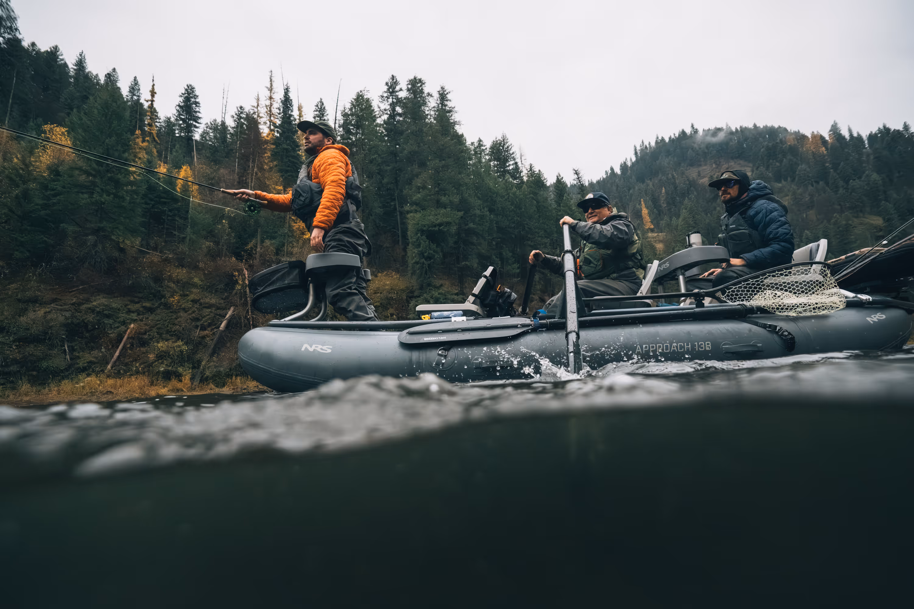 Man wearing sunglasses and outdoor gear fly fishing amid yellow flowering bushes under a cloudy sky.