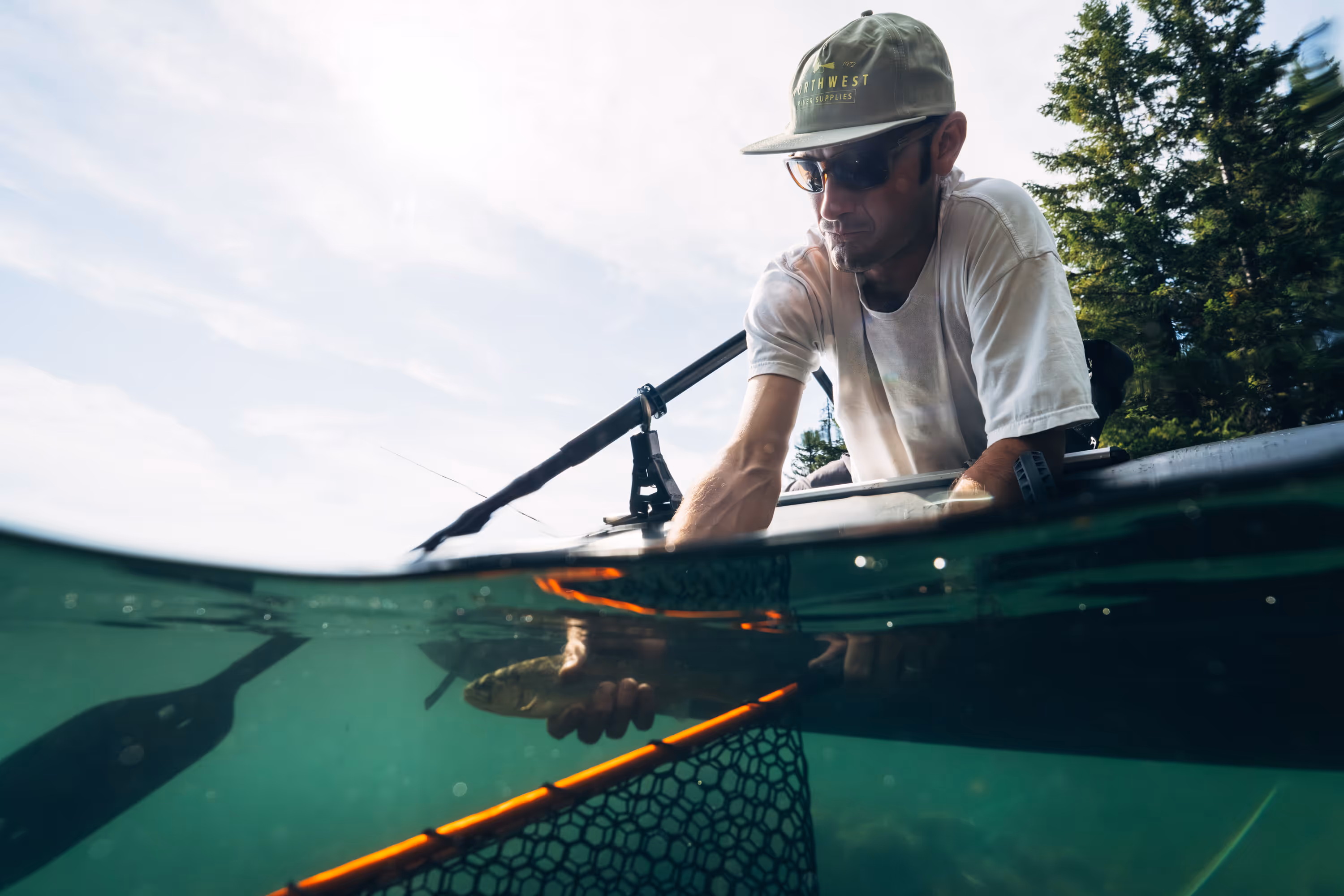 Man wearing sunglasses and a cap holding a fish over water while sitting in a kayak near green trees.