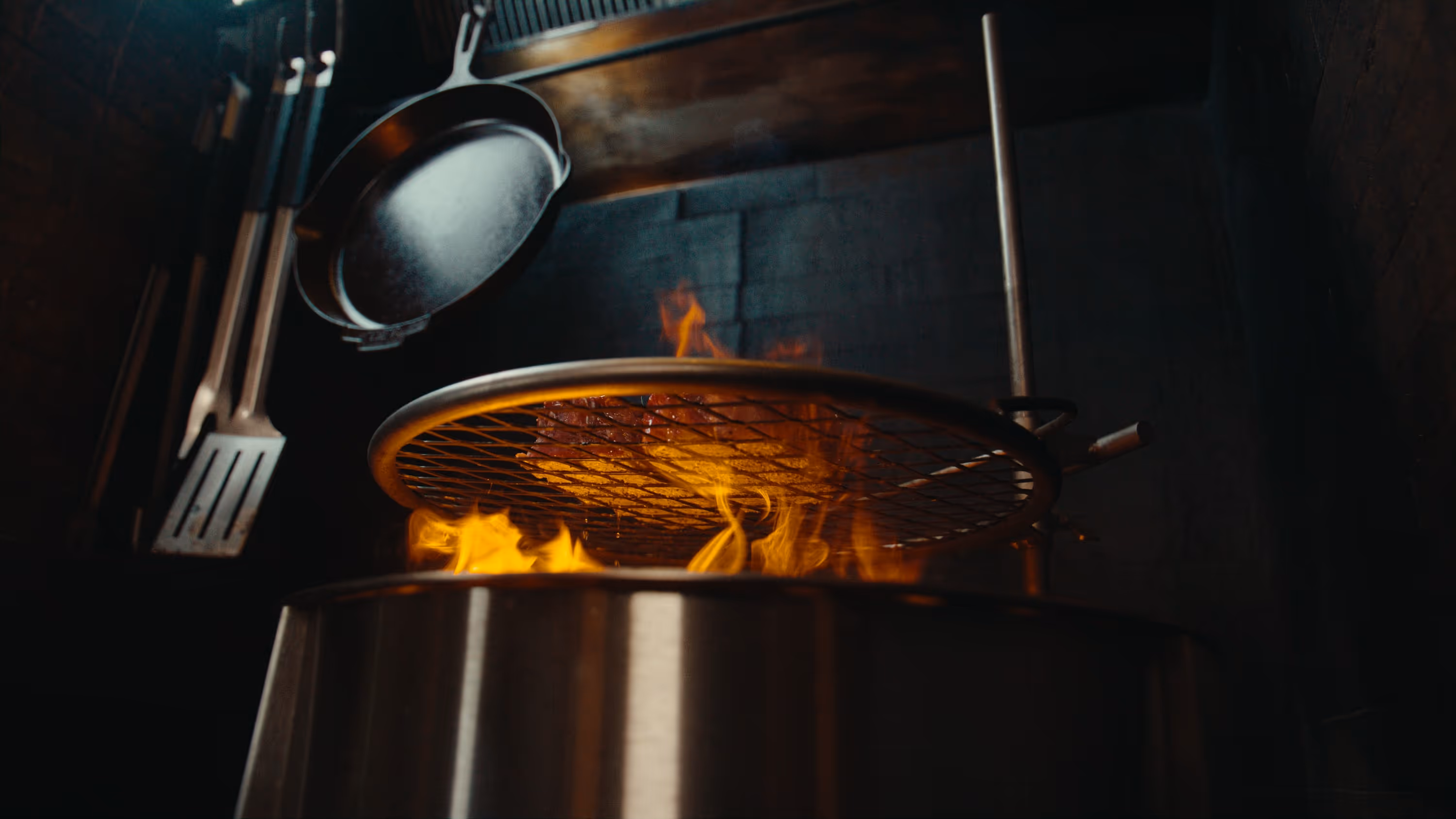 Meat grilling over an open flame on a round metal grate with kitchen utensils hanging nearby.