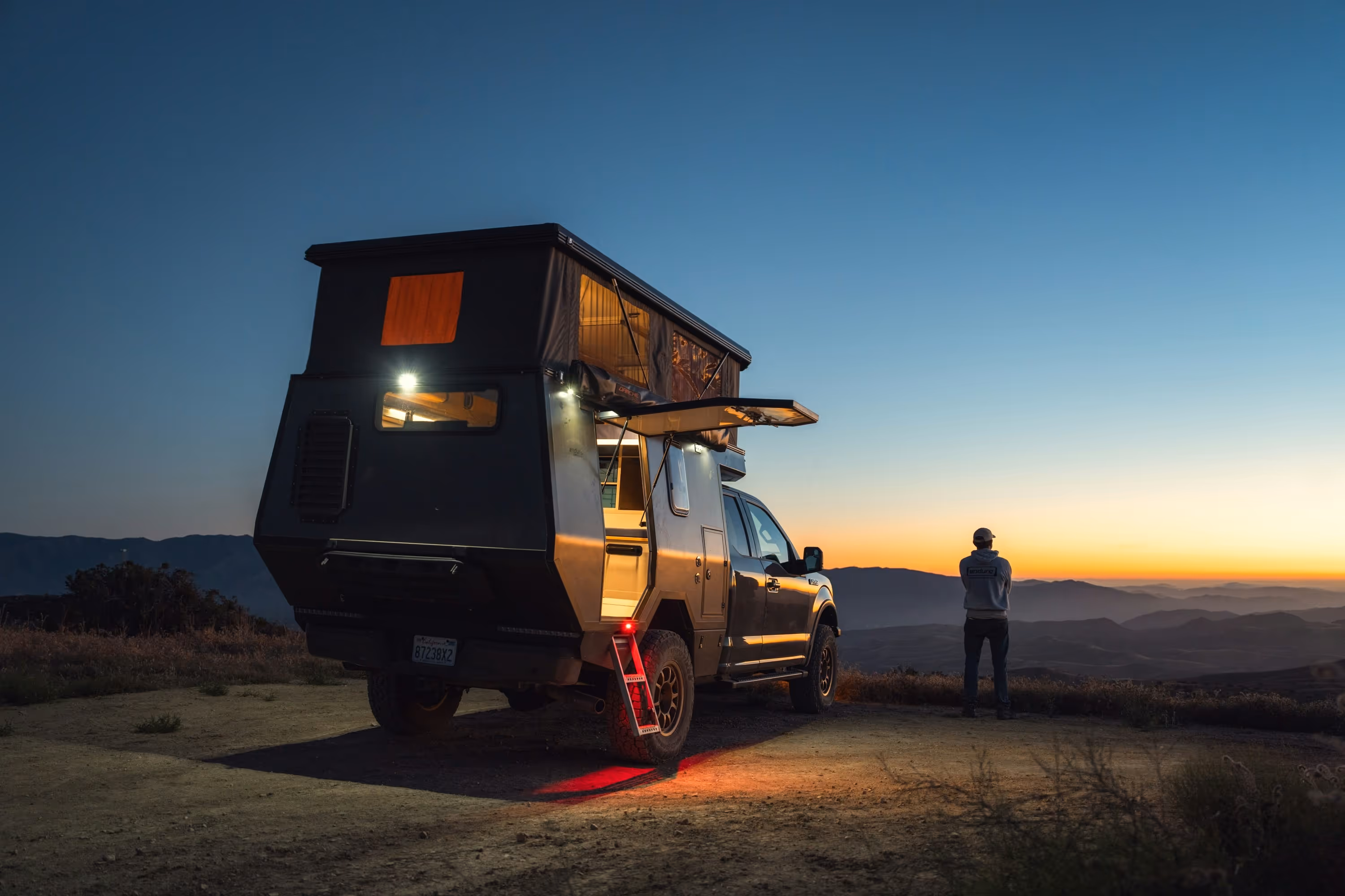 Pickup truck with rooftop camper parked on a hill at sunset with a person standing nearby overlooking mountains.