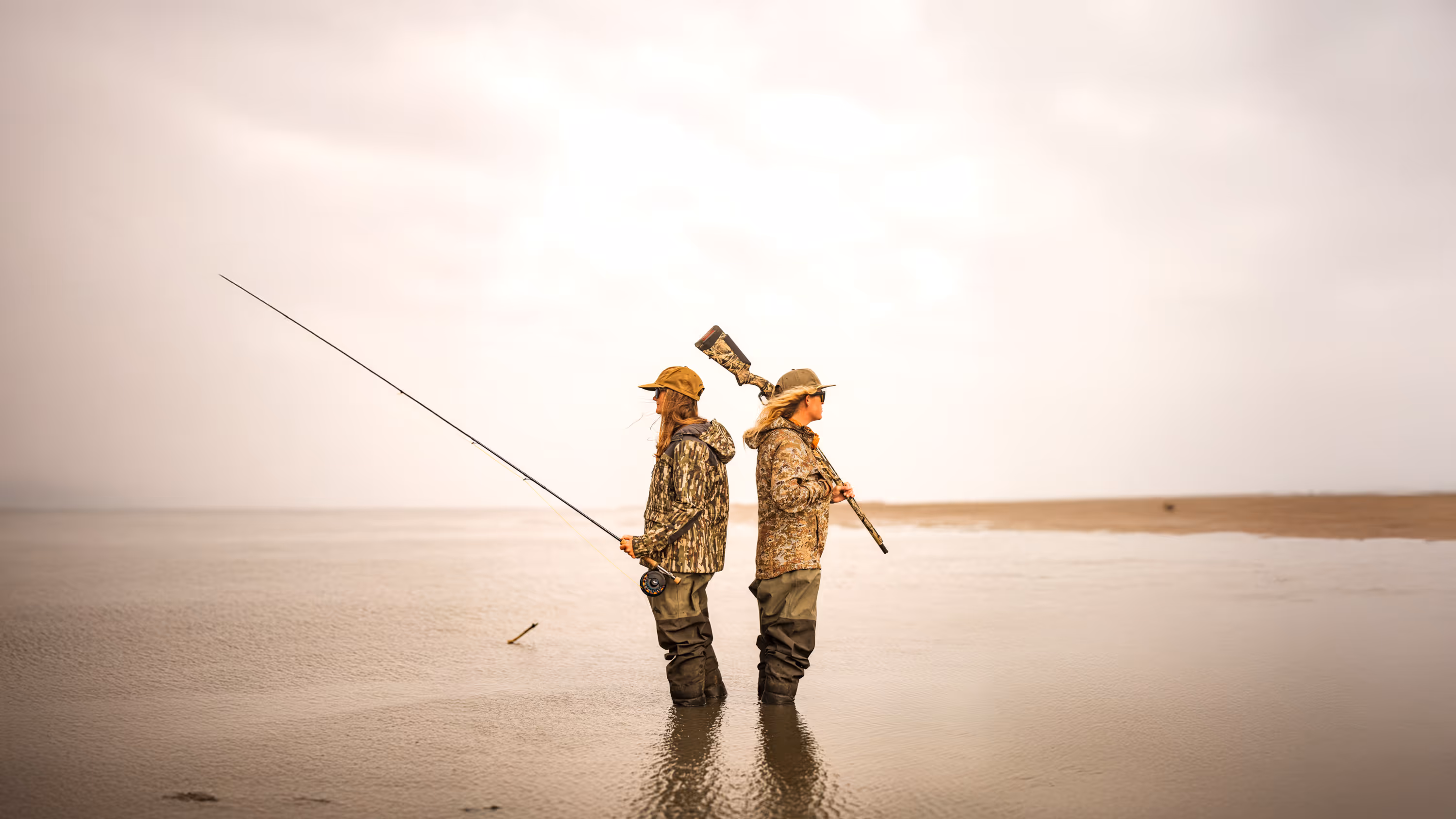 Two people wearing camouflage jackets and waders standing back-to-back in shallow water, one holding a fishing rod and the other a camouflaged rifle.