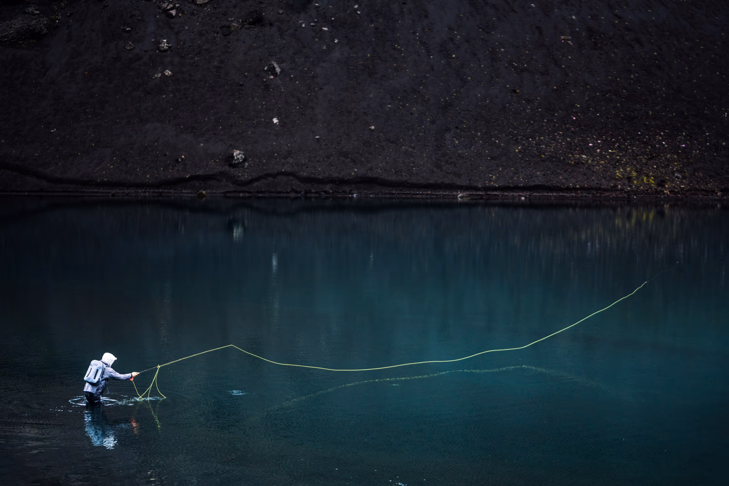 Person fly fishing in dark blue calm water near a rocky shore.