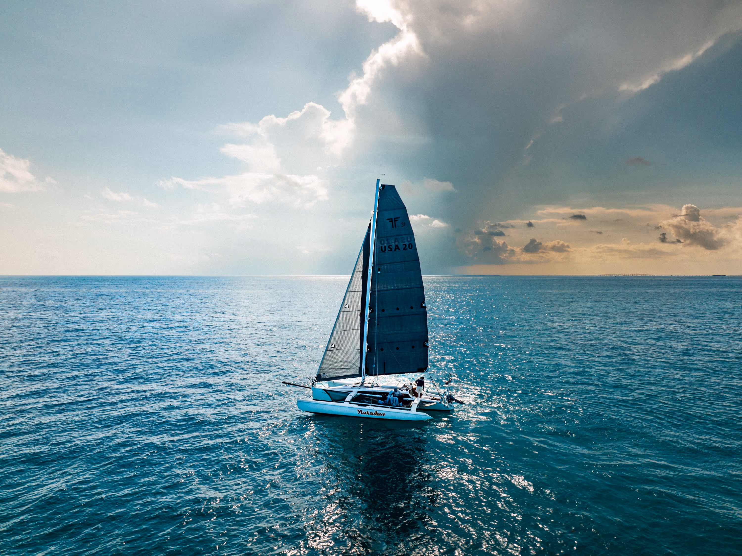 Sailboat named Matador with partially raised sails sailing on calm blue ocean under a sky with mixed clear and dark clouds.