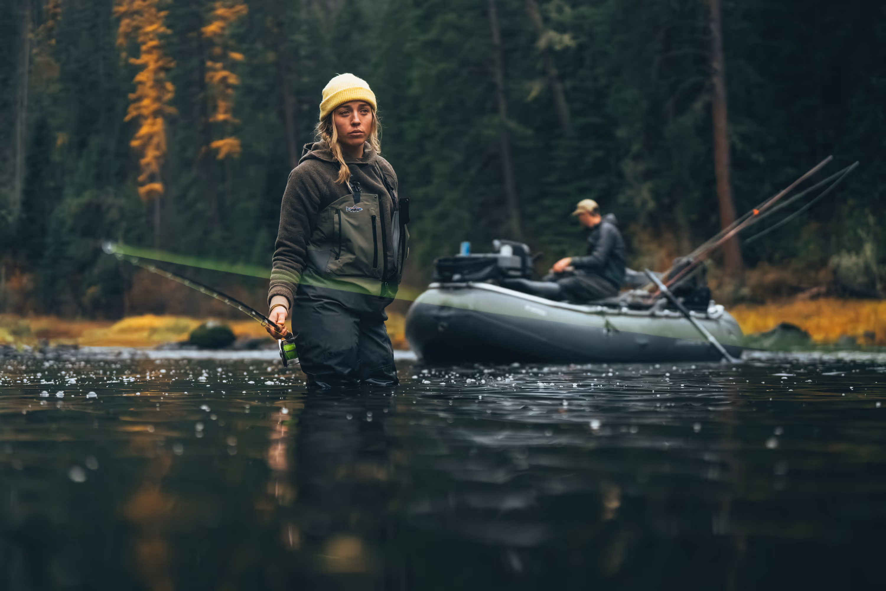 Person wearing outdoor gear with a blue backpack and brown pants walking on rocky terrain toward a blue helicopter hovering nearby.