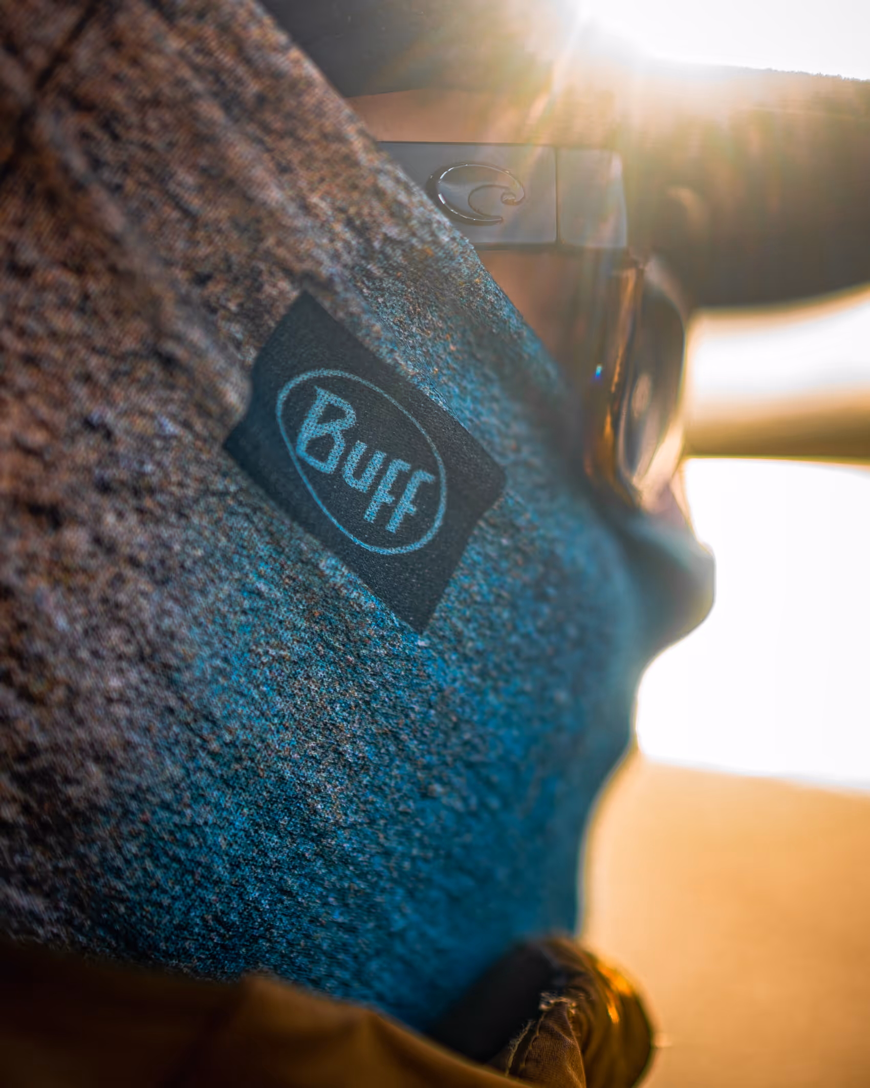 Close-up of a blue Buff brand neck gaiter worn with sunglasses in sunlight.