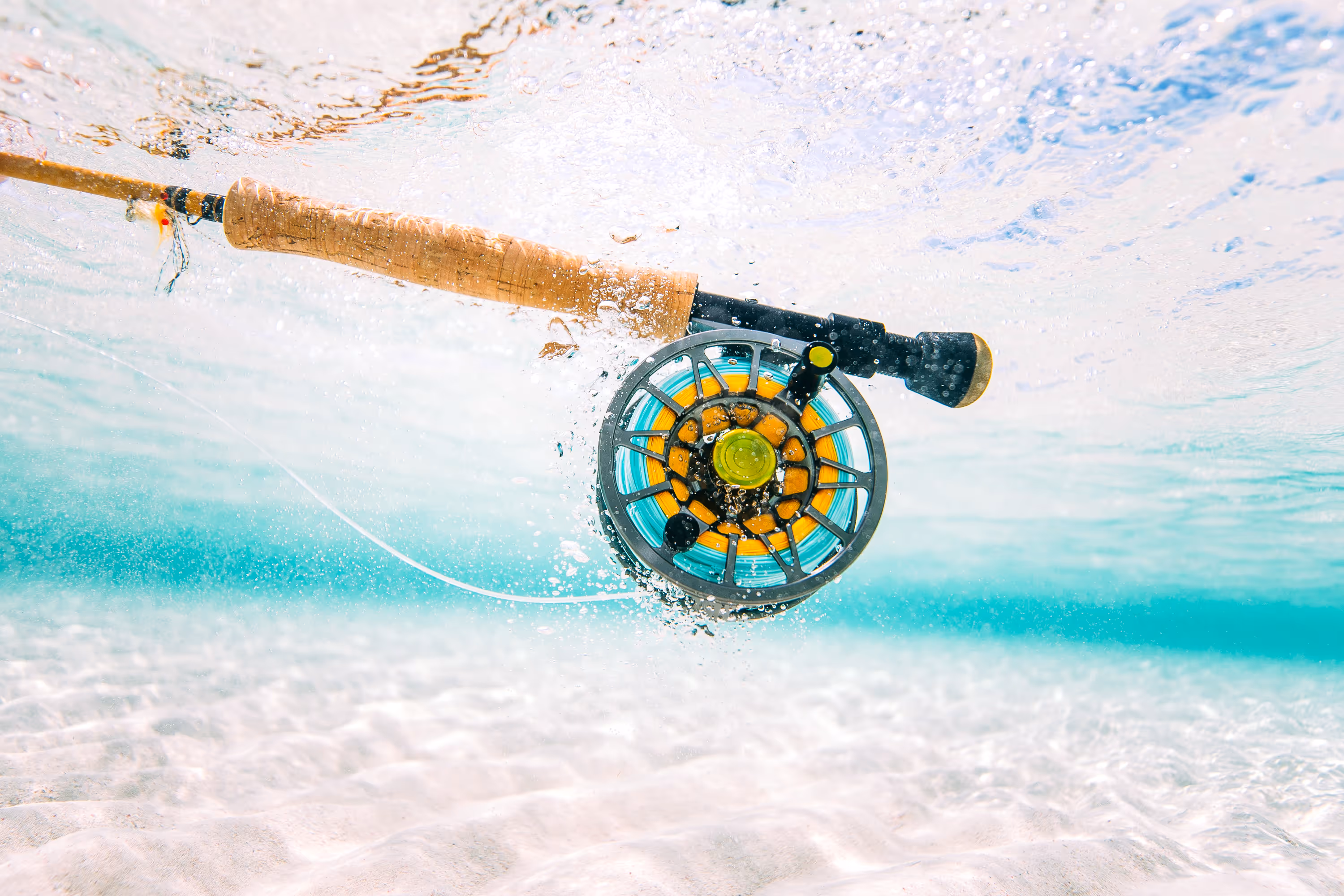 Close-up underwater view of a fly fishing rod and reel with clear turquoise water and sandy bottom.