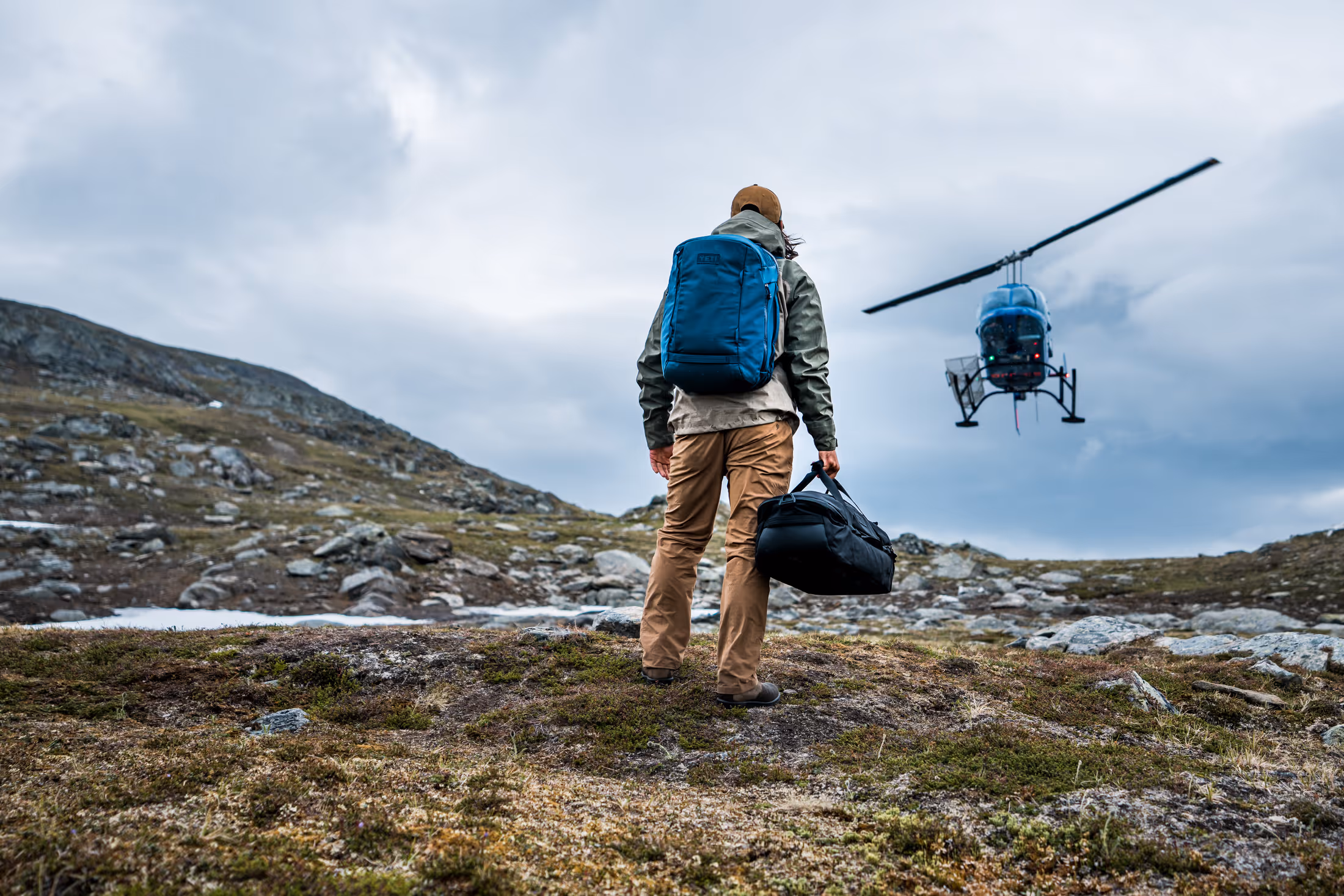 Person with a blue backpack and brown pants walking on rocky terrain toward a hovering blue helicopter.