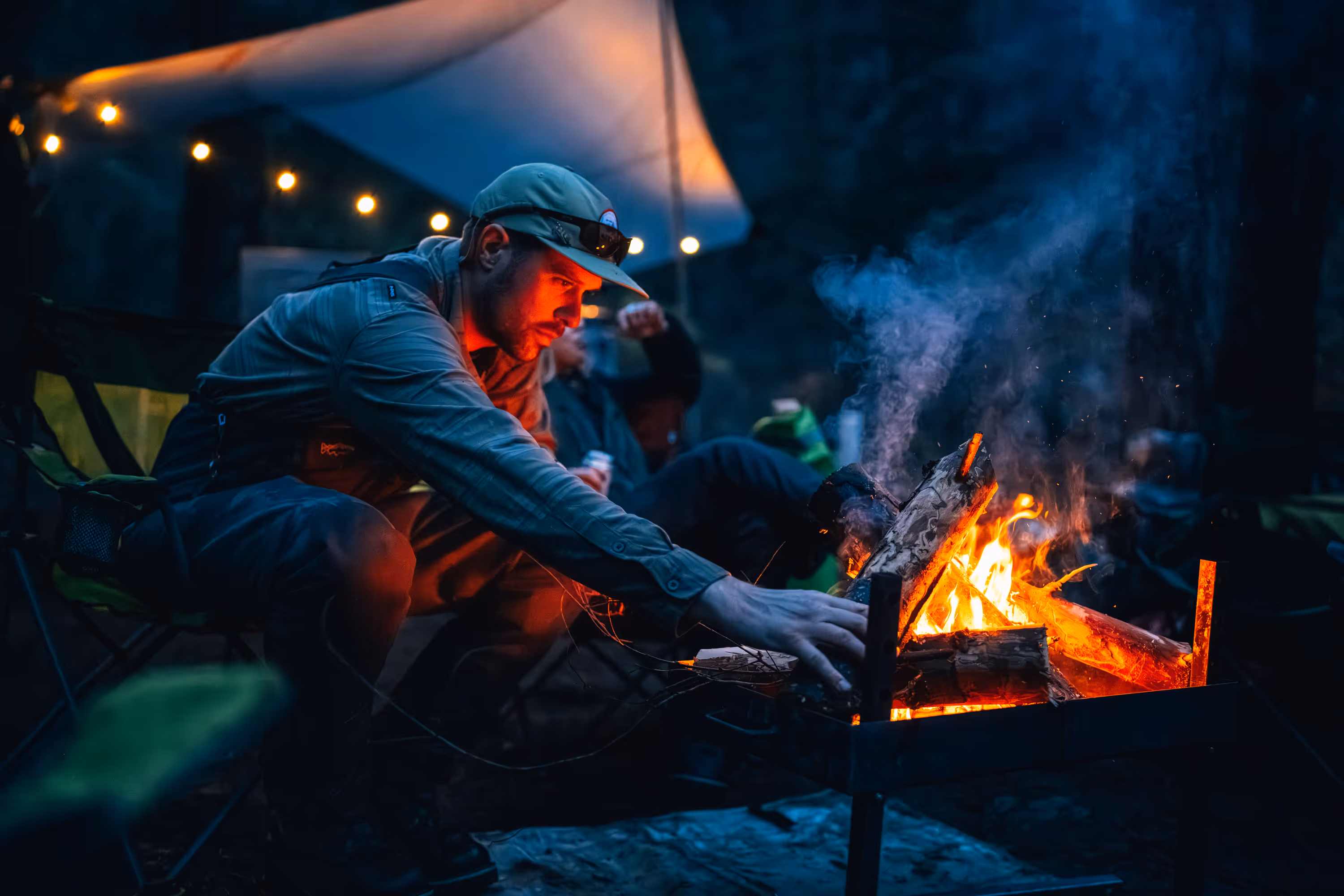 Man in outdoor clothing tending a campfire at night with string lights in the background.
