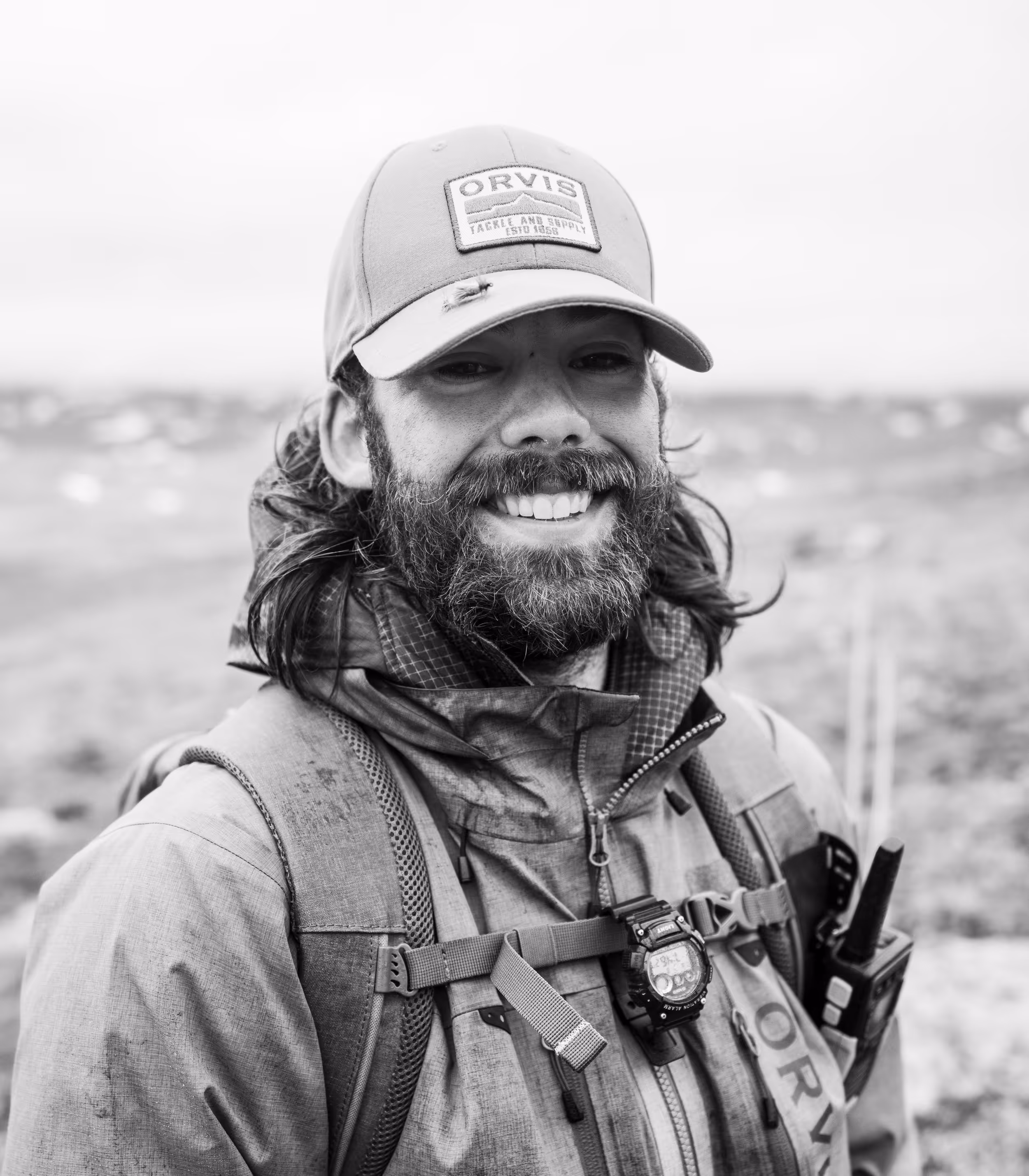 Man with long hair and beard wearing a white cowboy hat, sunglasses, and a blue-striped shirt leaning on a wooden fence with a green hillside and river in the background.