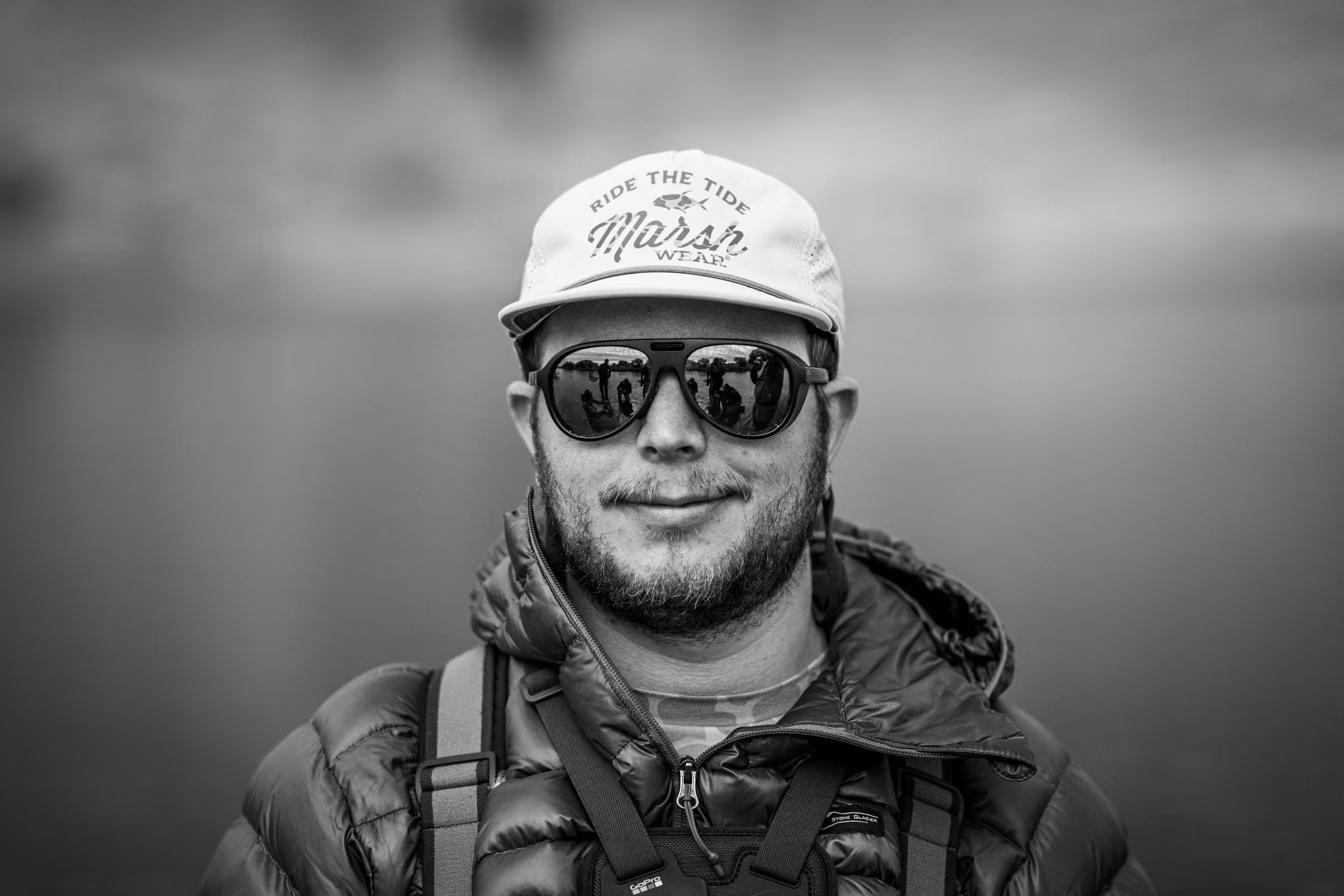 Smiling man wearing a rain jacket and backpack outdoors near a rocky river in a forest.