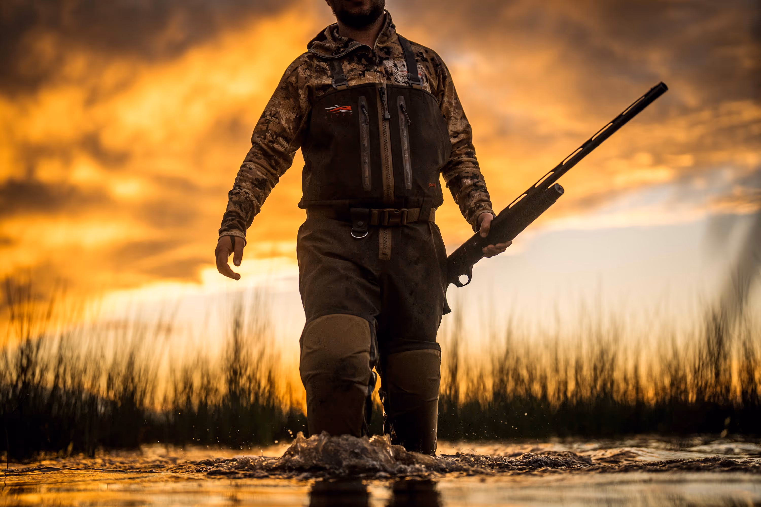 Person in camouflage and waders holding a shotgun walking through water during sunset.
