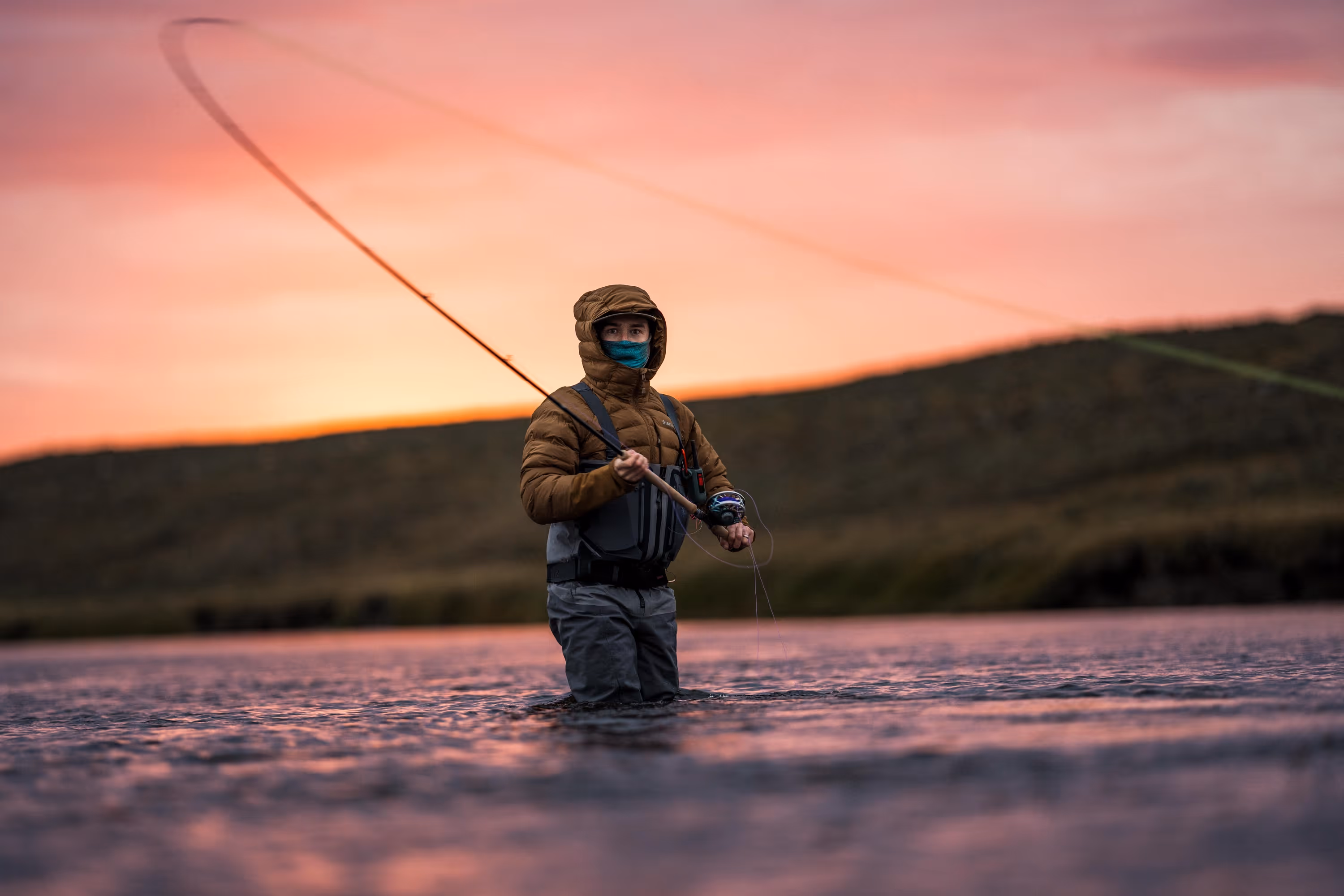 Person wearing a hooded jacket and face mask fly fishing in a river at sunset with hills in the background.