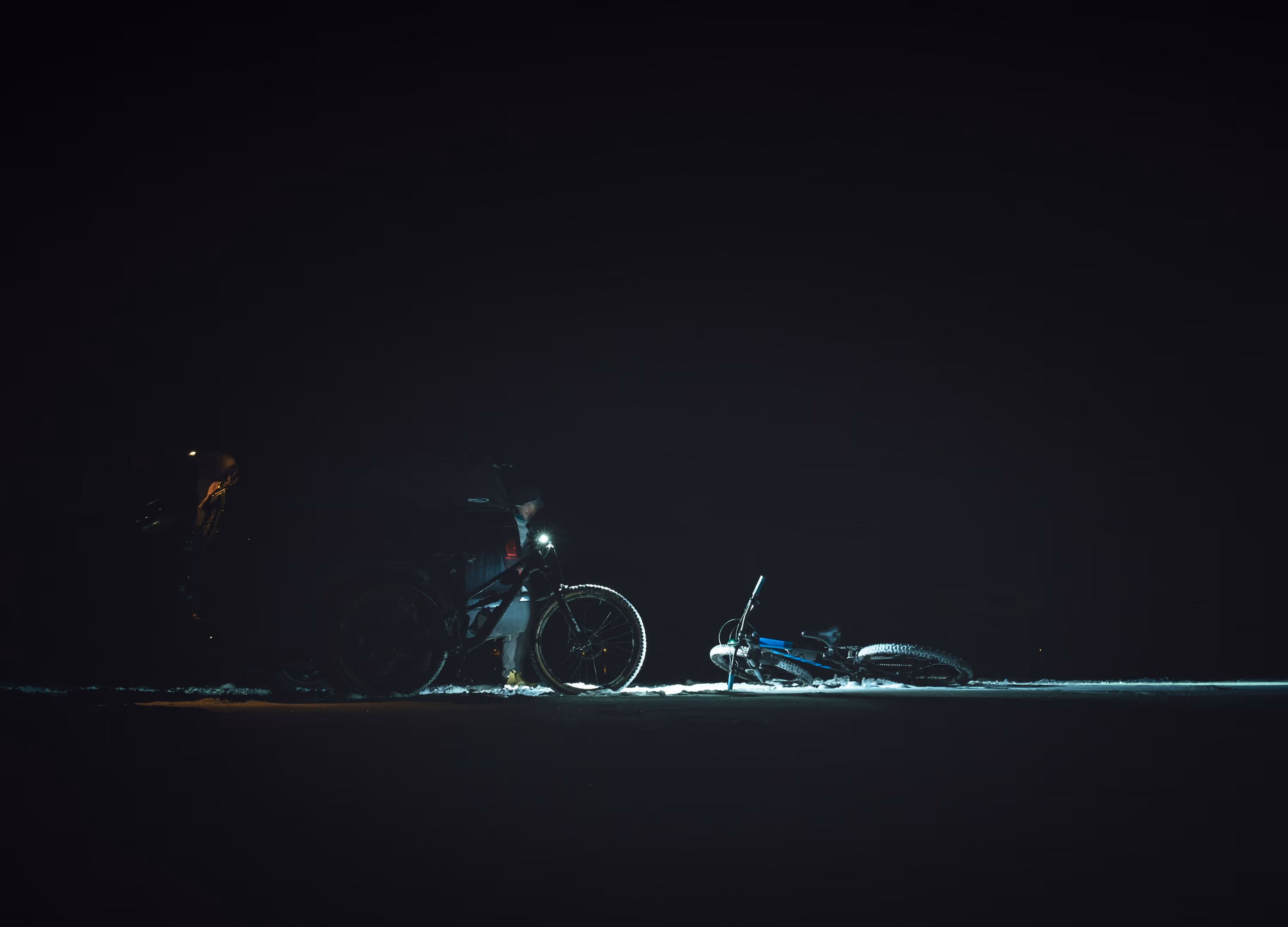 Person standing next to a mountain bike with a headlight shining on a fallen bicycle on snow at night.