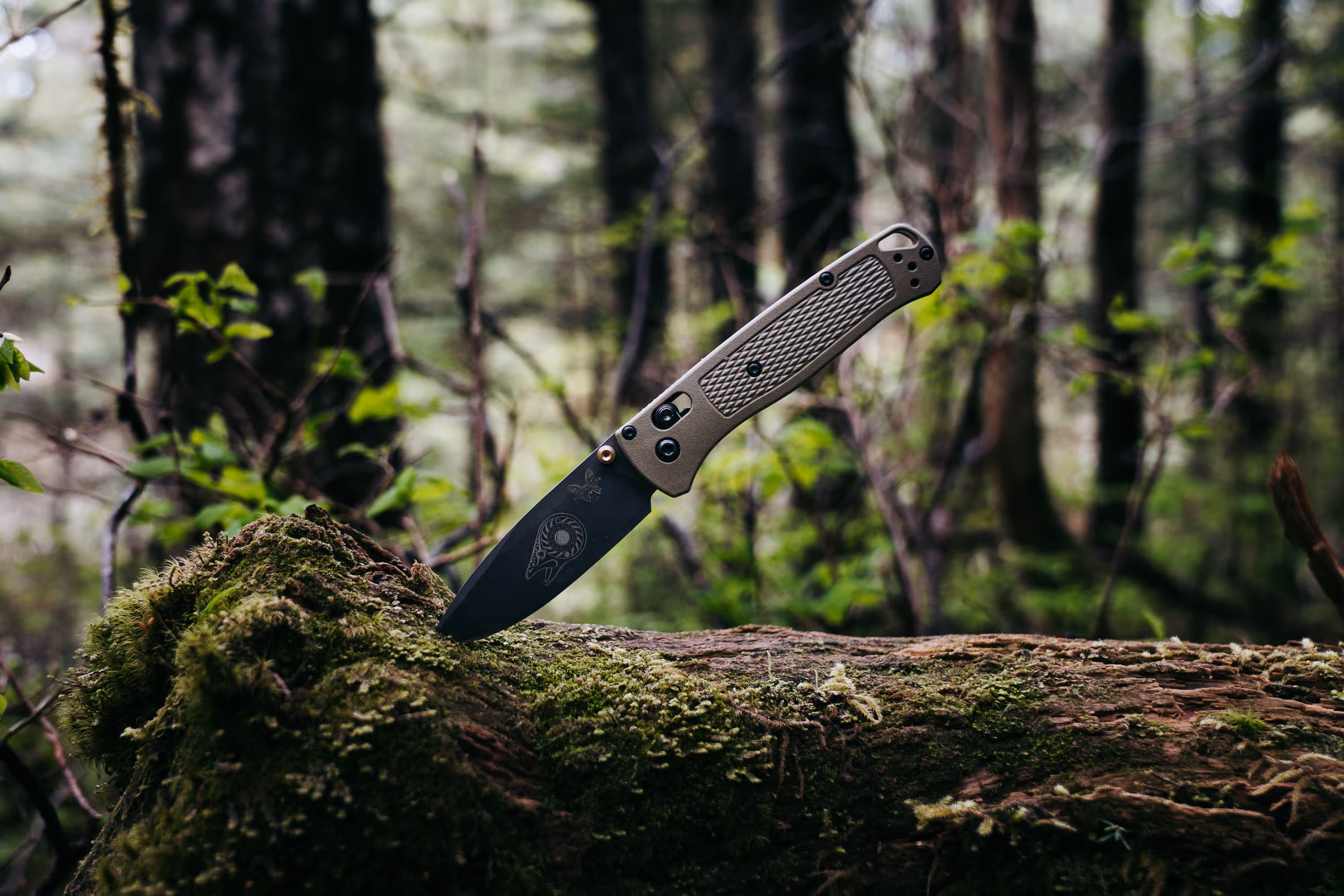 Folding knife with engraved blade stuck into moss-covered log in a forest setting.