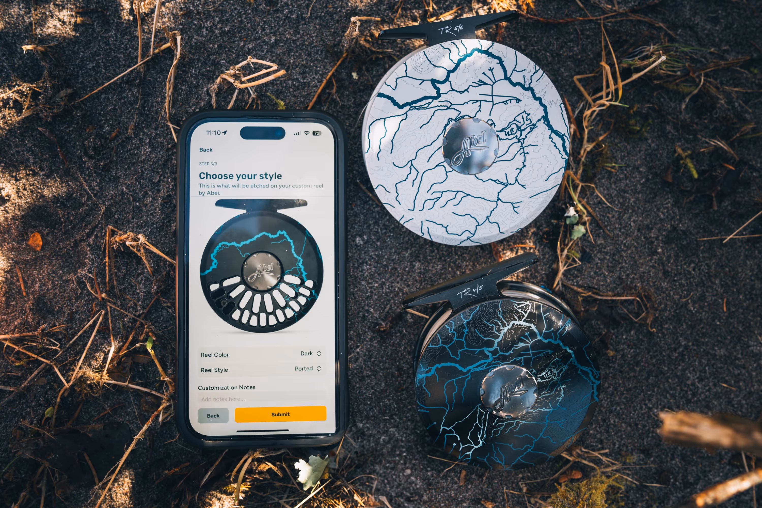 Smartphone displaying a custom reel style selection screen next to two fishing reels with intricate river-like patterns on a sandy surface.