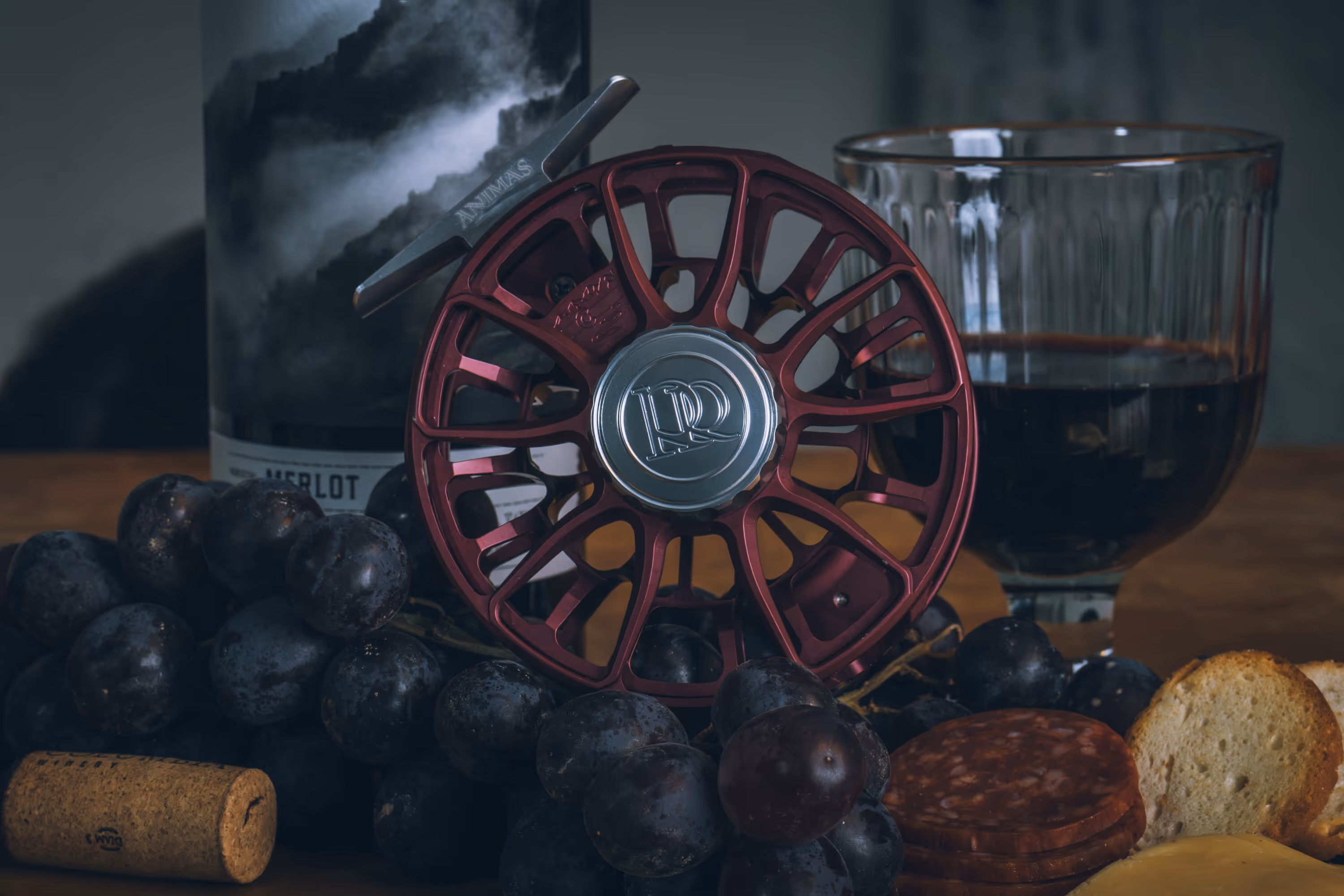 Red fishing reel surrounded by dark grapes, sliced salami, cheese, bread, a wine bottle, and a glass of red wine.
