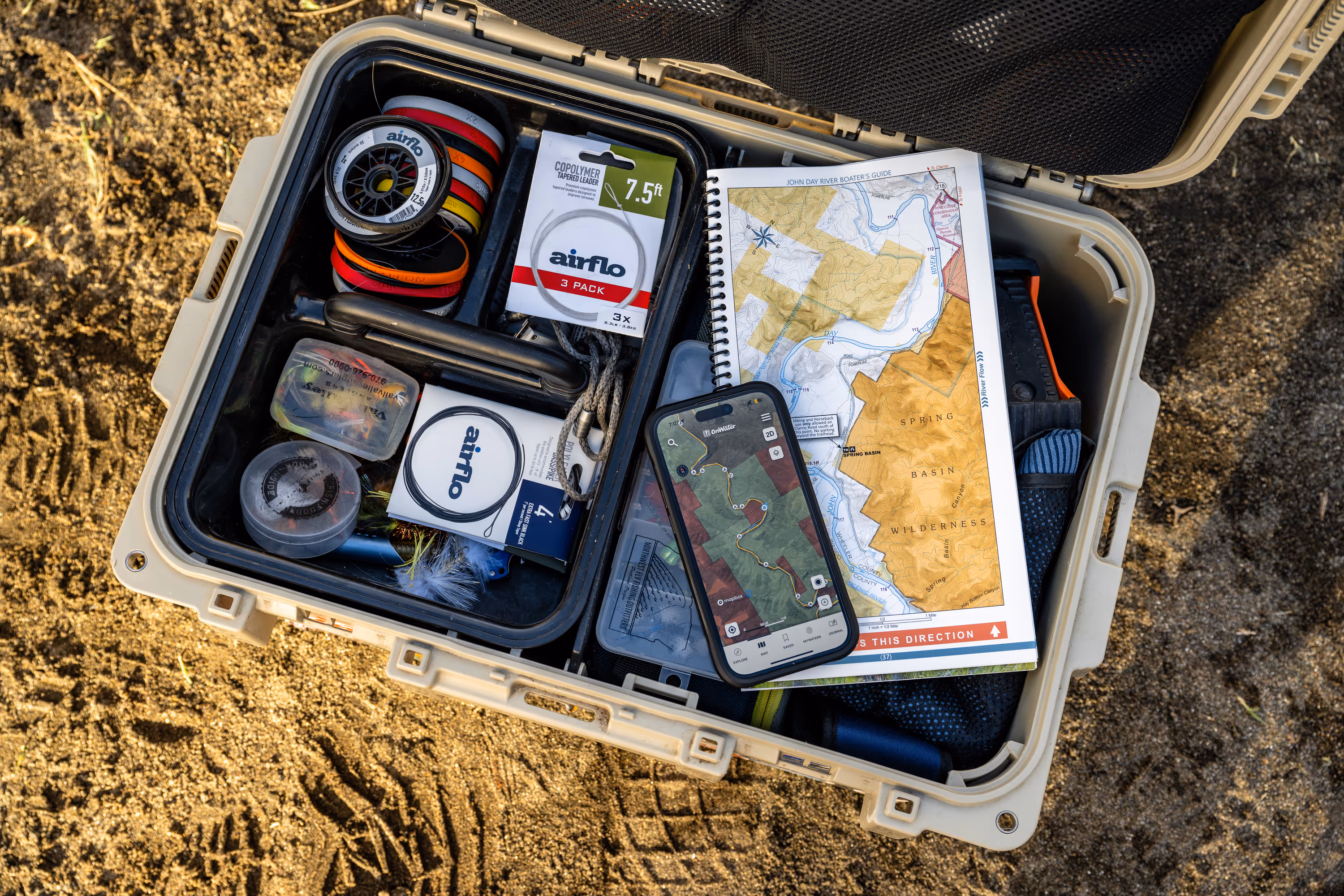 Open tan tackle box on sandy ground containing fishing flies, airflo brand fishing line, a river map, and a smartphone displaying a river navigation app.