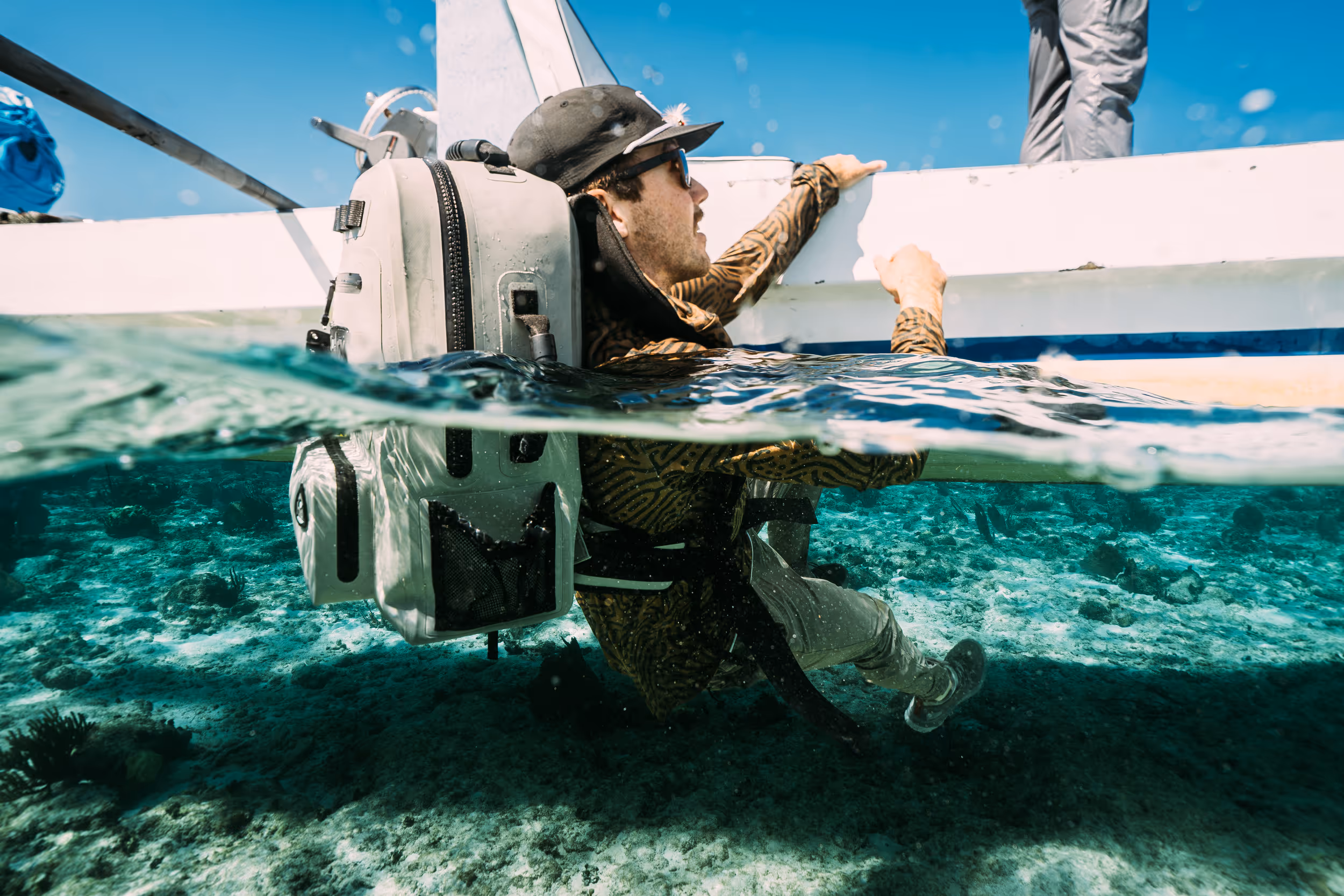 Man wearing a cap and sunglasses climbing onto a boat from clear blue water, carrying a waterproof backpack.