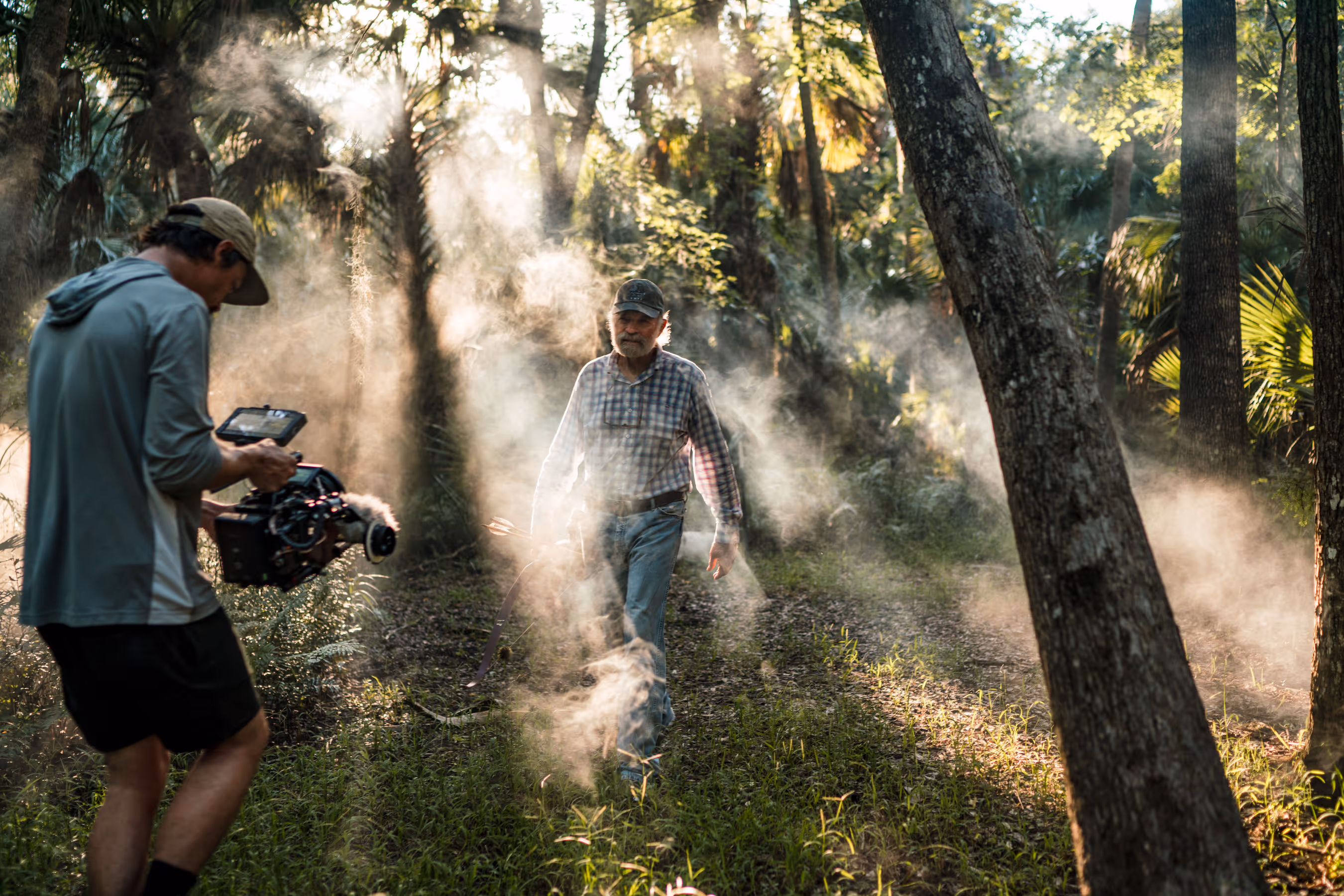 Elderly man with a beard wearing a plaid shirt, jeans, and a baseball cap walks in a misty forest holding arrows and a bow.
