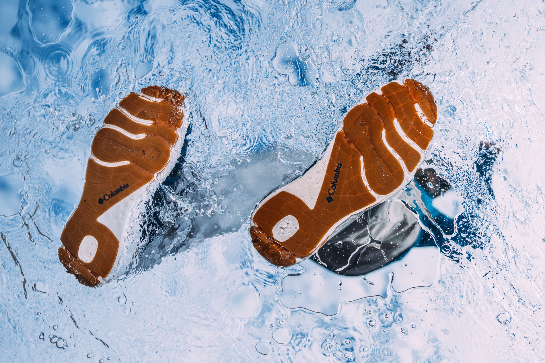 Soles of brown and white Columbia brand shoes submerged underwater with bubbles and ripples.