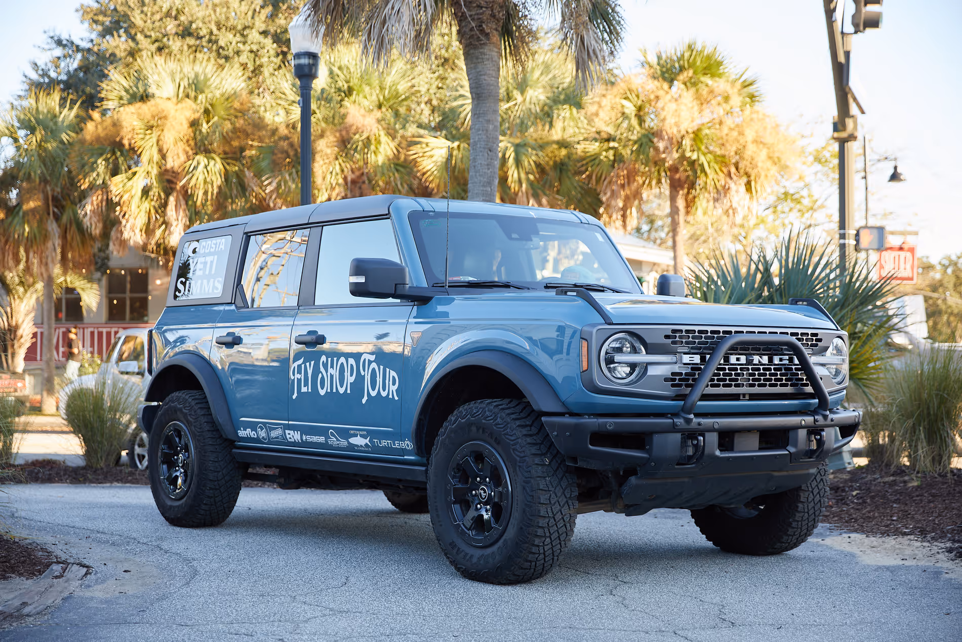 Blue Ford Bronco SUV with 'Fly Shop Tour' and brand logos parked on a street with palm trees in the background.
