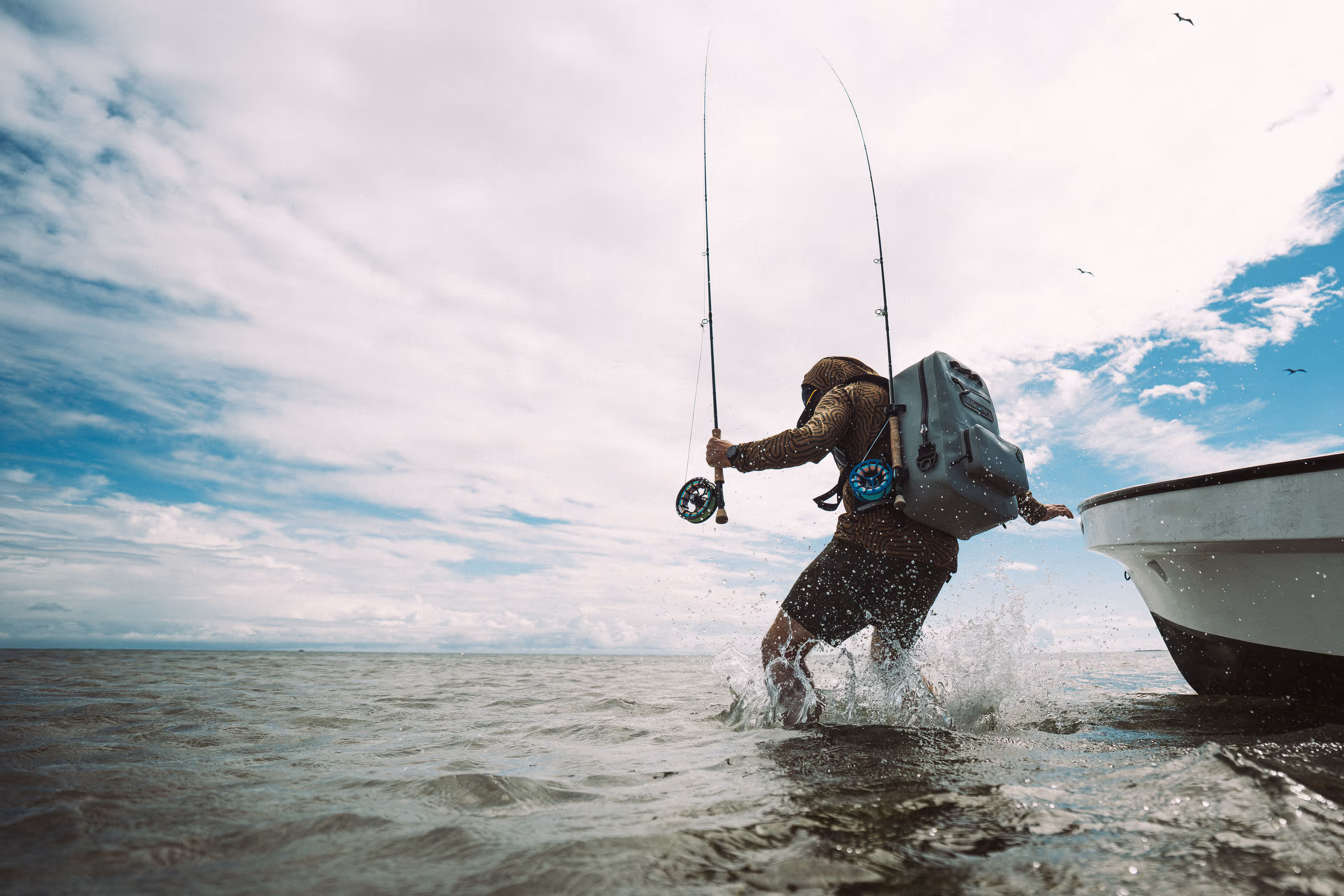 Person wearing a hood and backpack stepping out of a boat into shallow water, holding two fishing rods with splashing water around.
