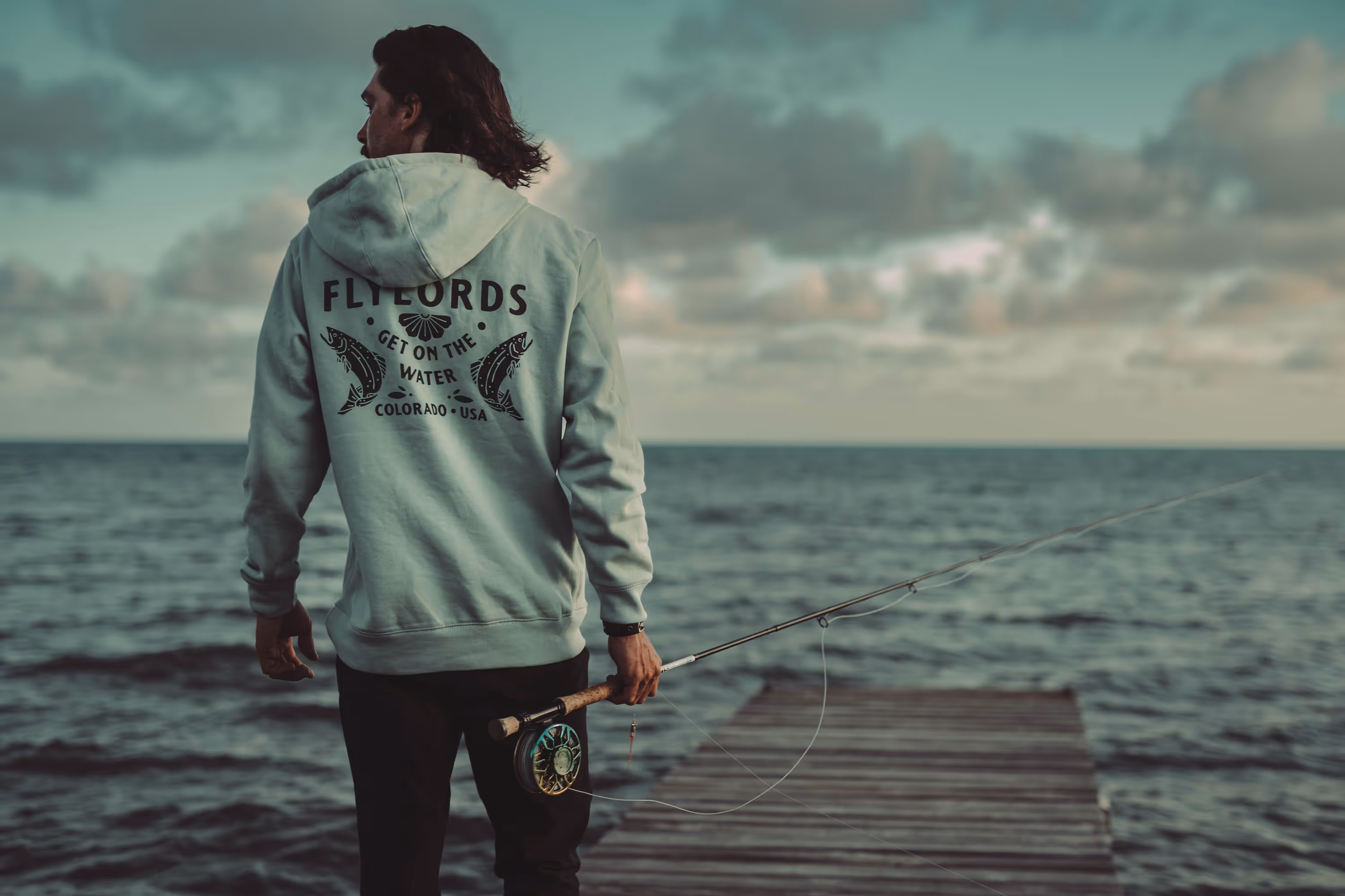 Person wearing a hoodie with fishing graphics holding a fishing rod and standing on a wooden dock over water at sunset.
