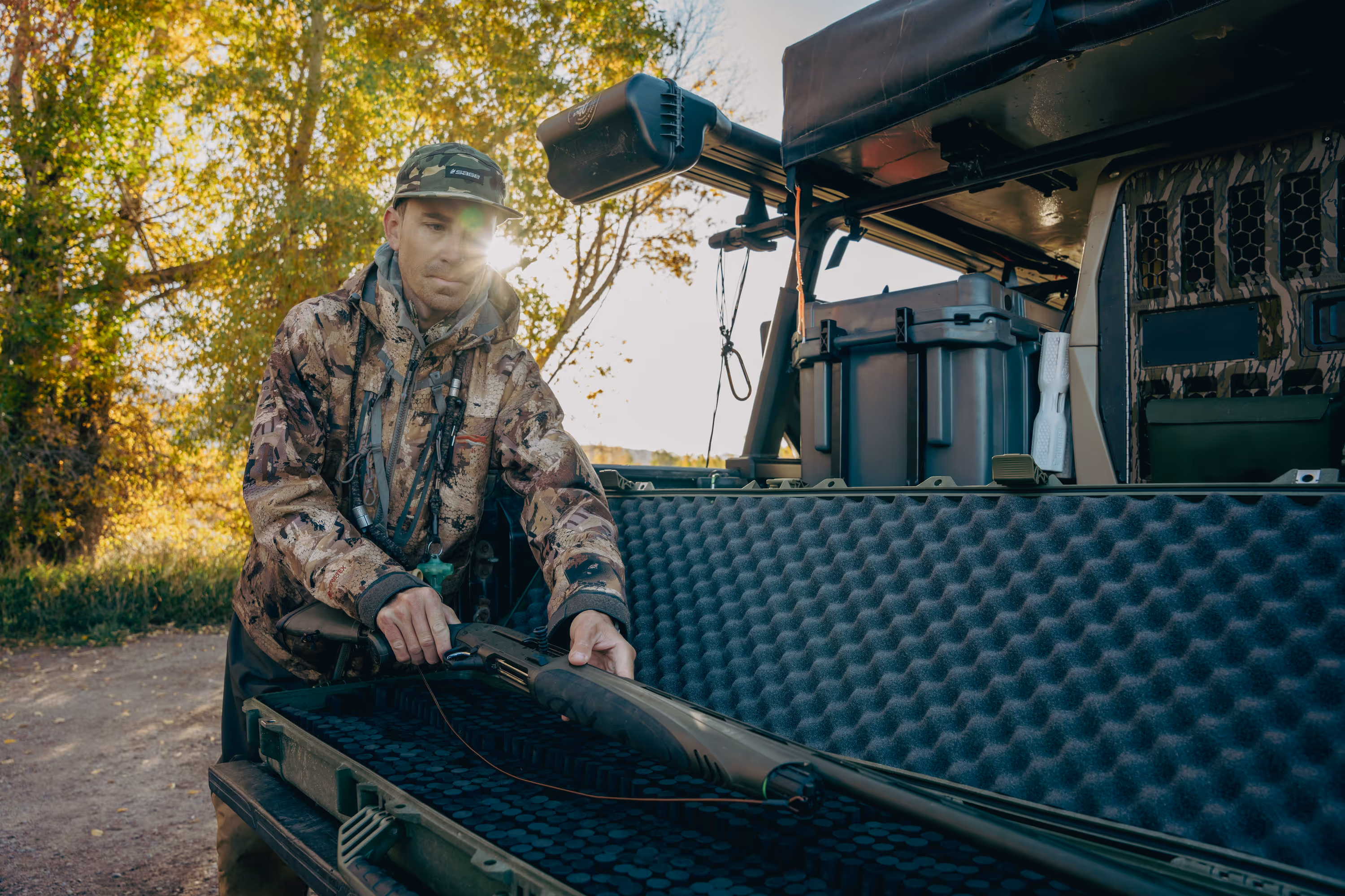 Two people wearing camouflage jackets and waders standing back-to-back in shallow water, one holding a fishing rod and the other a camouflaged rifle.