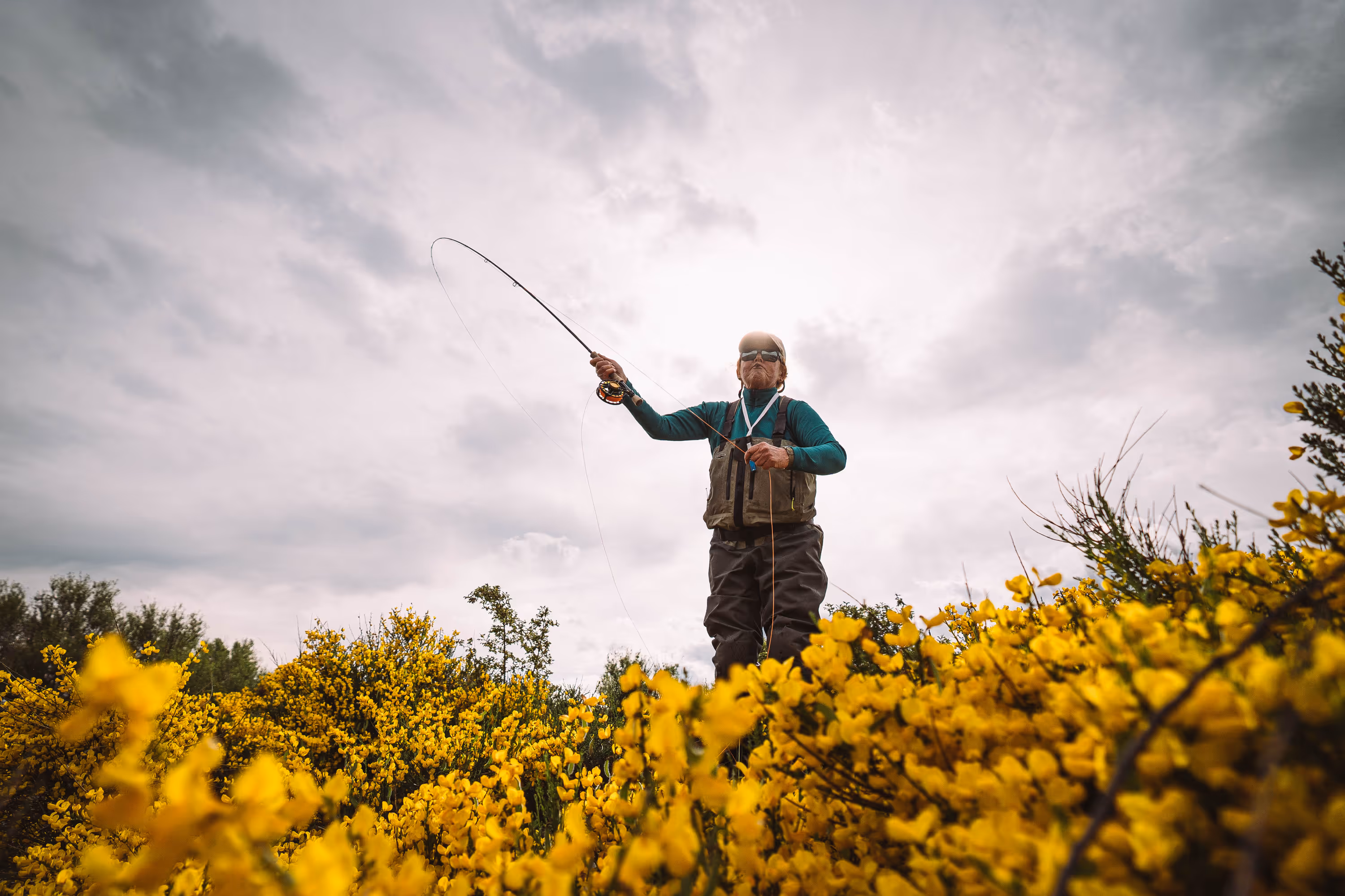 Two people wearing camouflage jackets and waders standing back-to-back in shallow water, one holding a fishing rod and the other a camouflaged rifle.