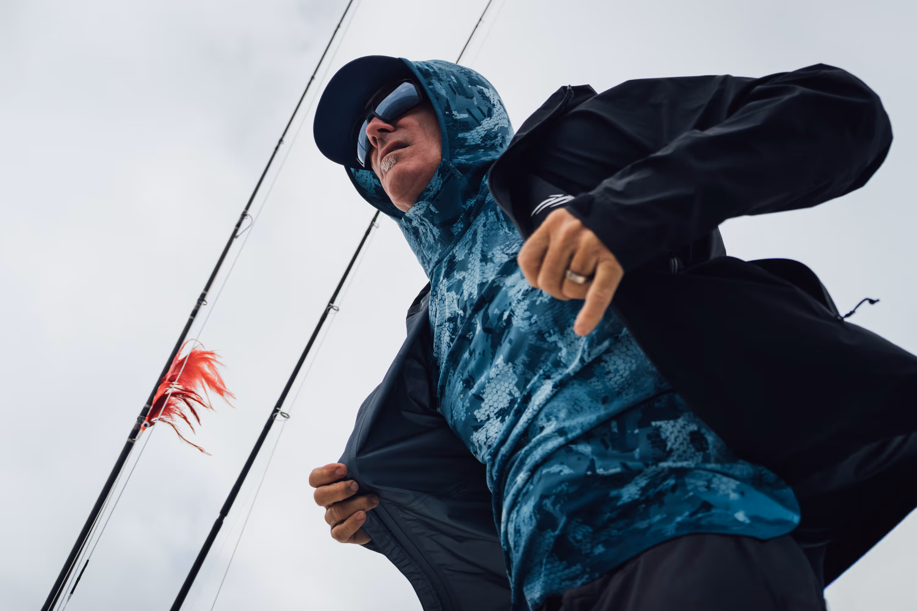 Two people wearing camouflage jackets and waders standing back-to-back in shallow water, one holding a fishing rod and the other a camouflaged rifle.