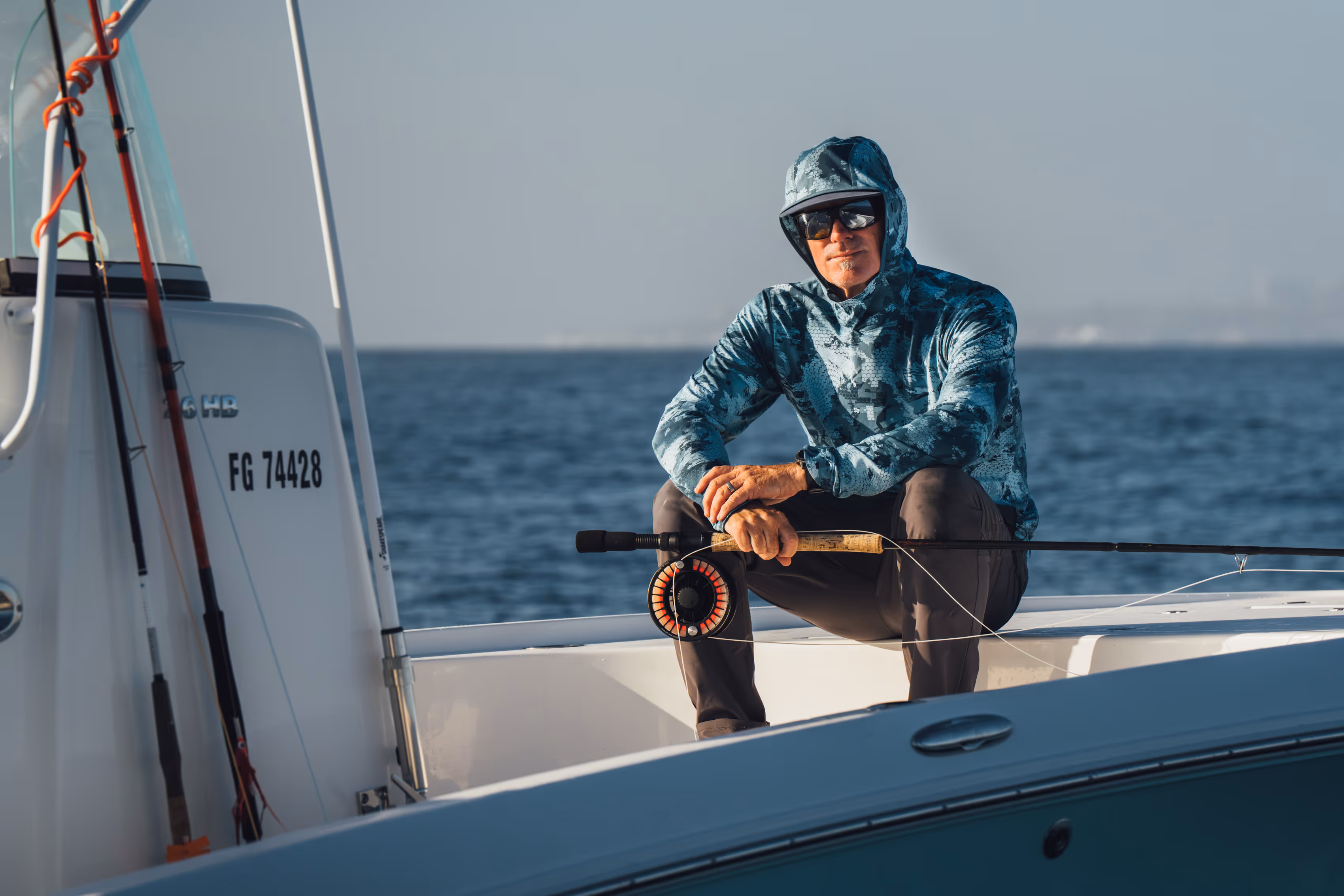 Two people wearing camouflage jackets and waders standing back-to-back in shallow water, one holding a fishing rod and the other a camouflaged rifle.