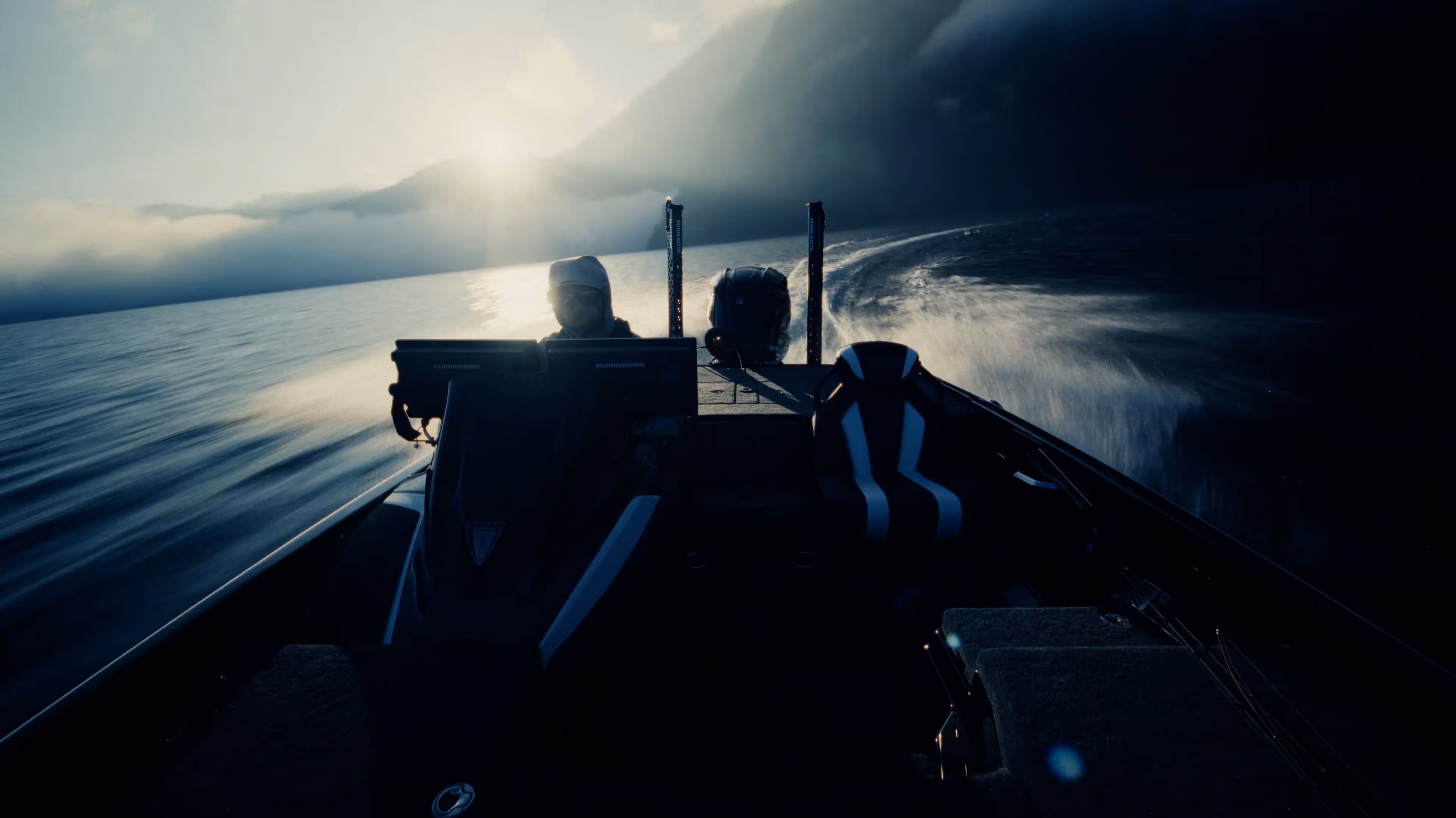Two people wearing camouflage jackets and waders standing back-to-back in shallow water, one holding a fishing rod and the other a camouflaged rifle.
