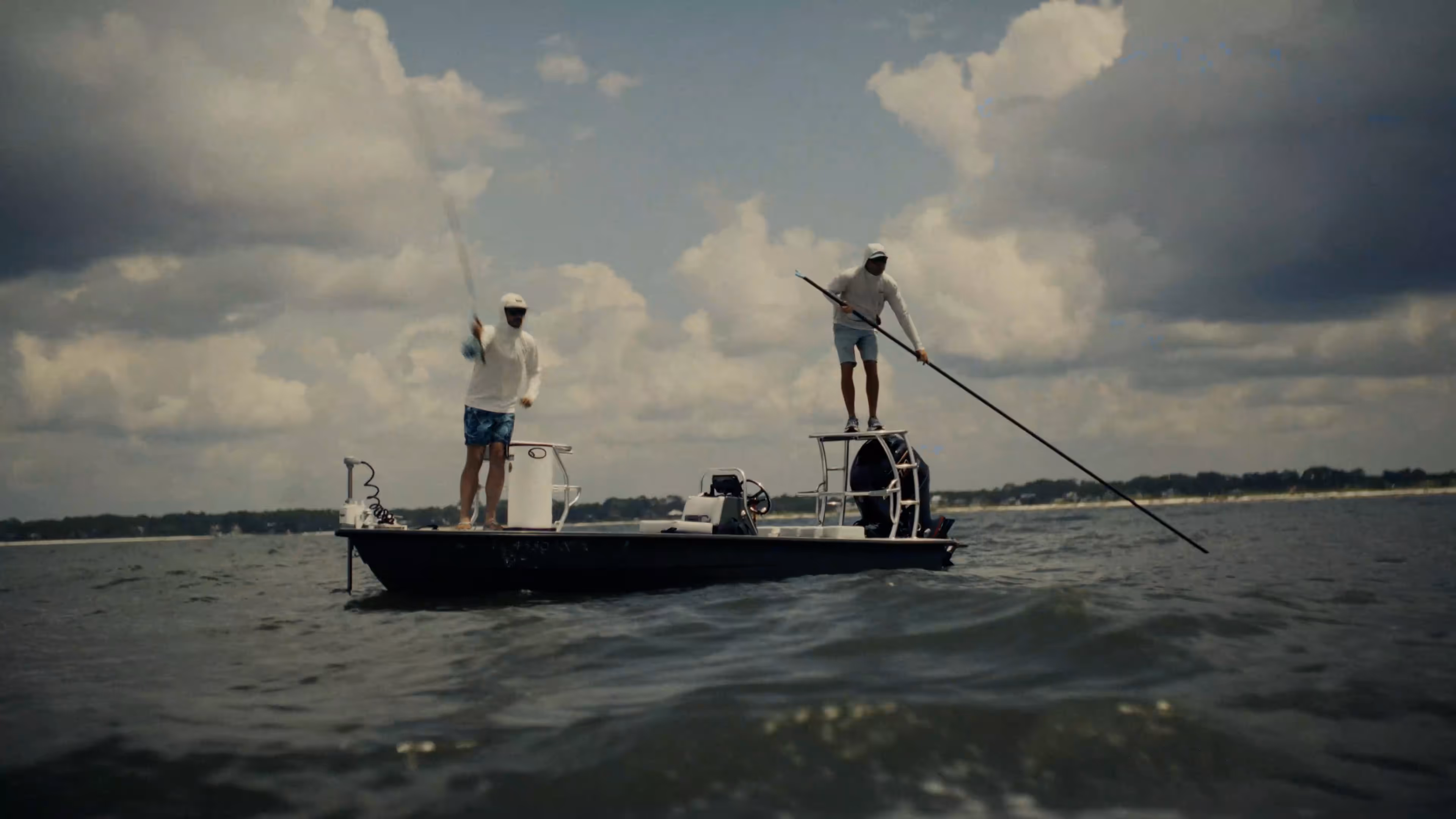 Two people wearing camouflage jackets and waders standing back-to-back in shallow water, one holding a fishing rod and the other a camouflaged rifle.