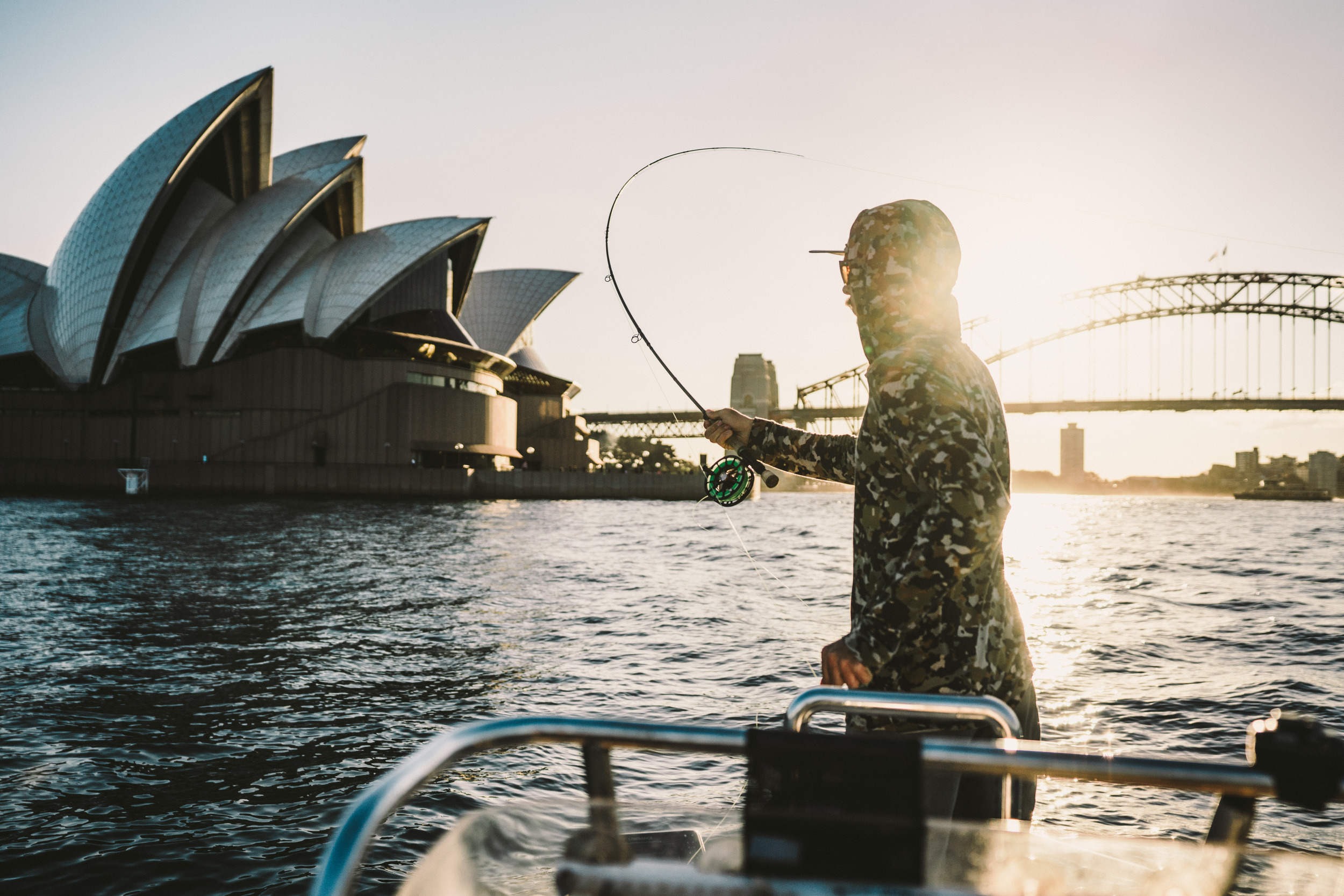 Person wearing a hood and backpack stepping out of a boat into shallow water, holding two fishing rods with splashing water around.